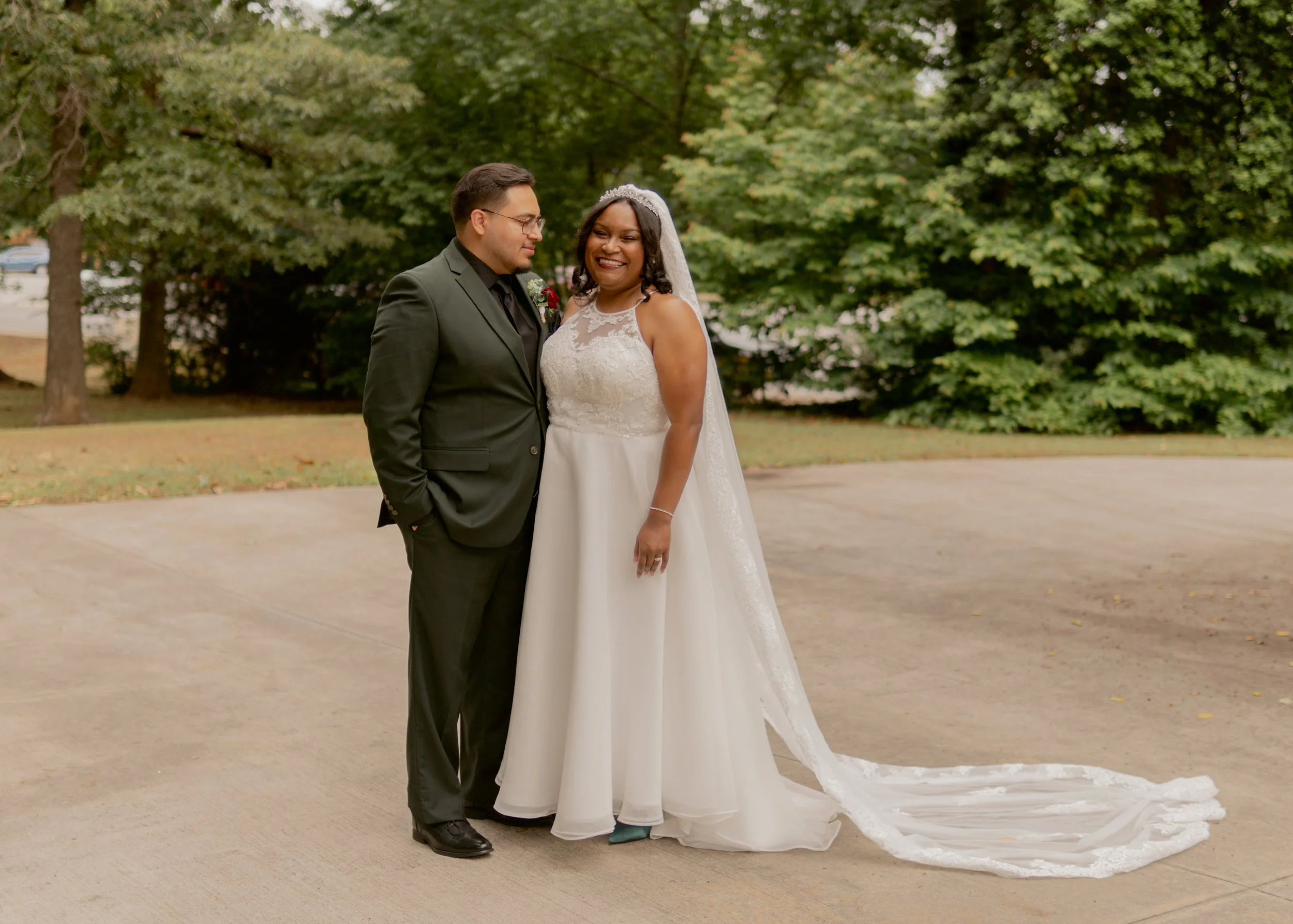 A smiling bride and groom in wedding attire standing outdoors on a pathway, with trees in the background.