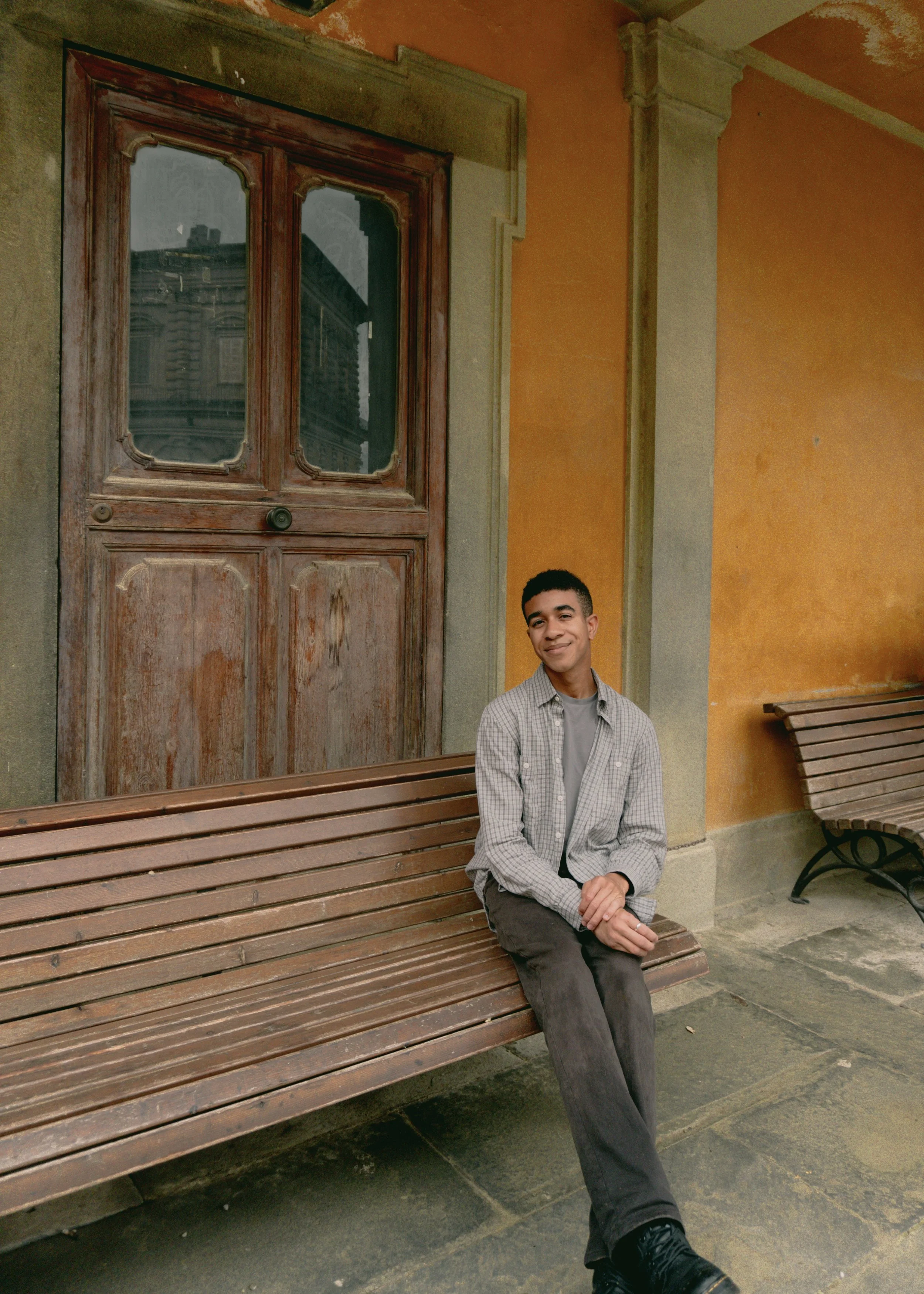 A young man with short dark hair and light brown skin sitting on a wooden park bench. He is wearing a light grey plaid shirt, grey t-shirt, dark grey pants, and black shoes. Behind him is a yellow-orange wall with an ornate wooden window reflecting buildings.