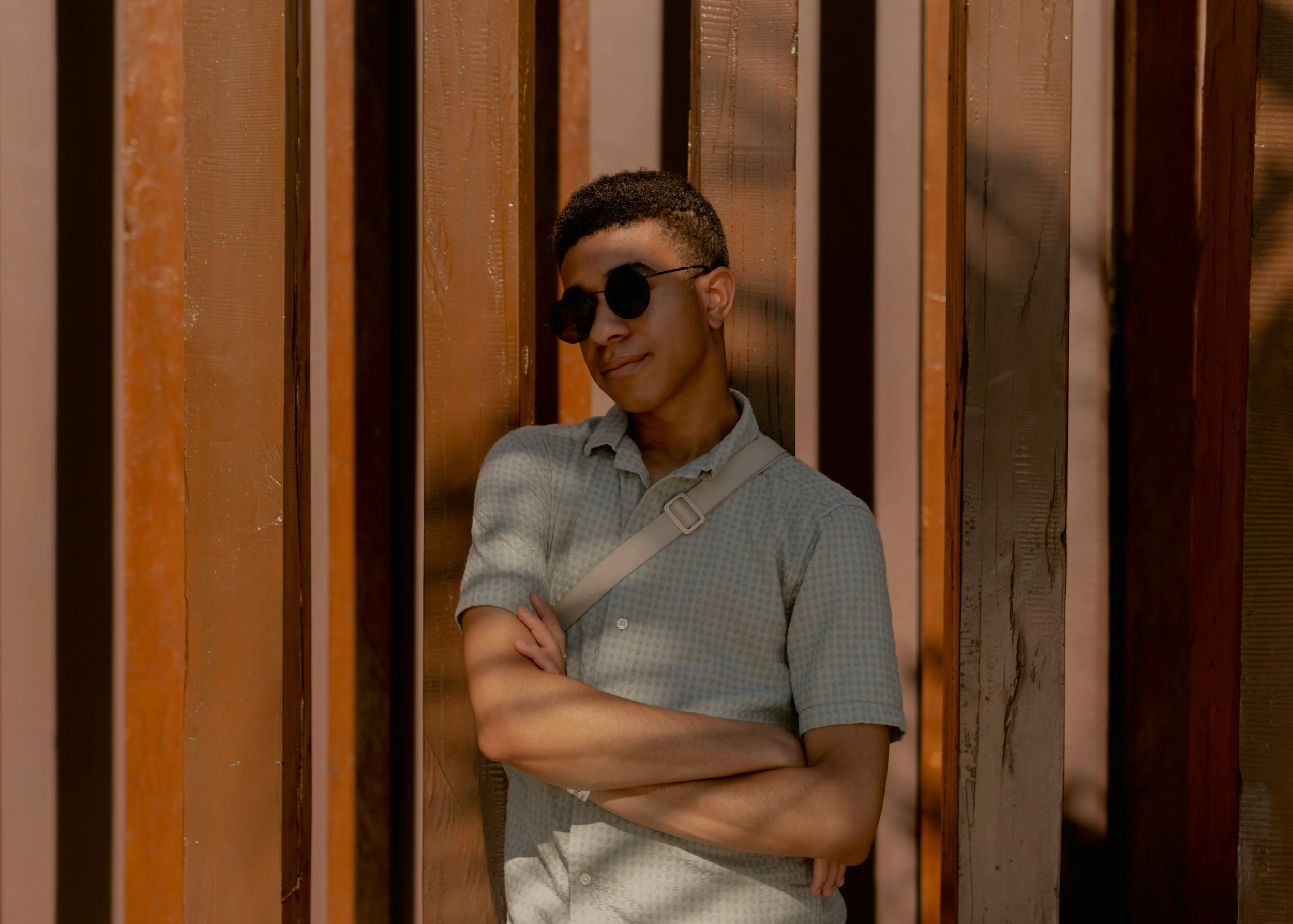 A young man with short curly hair wearing sunglasses and a checked shirt, standing with his arms crossed, leaning against a wooden wall with vertical slats.