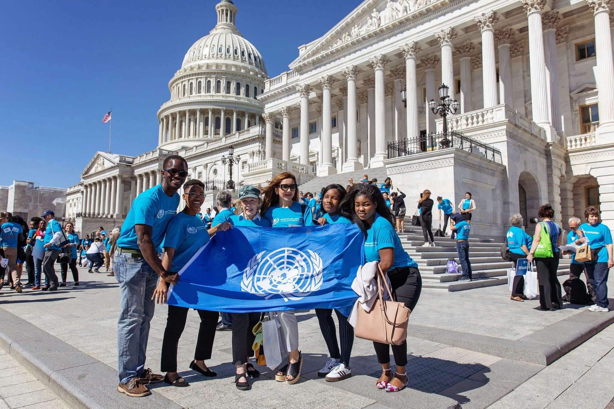 A diverse group of people holding a United Nations flag, standing in front of the U.S. Capitol building during a sunny day with a clear blue sky. Many are wearing blue shirts. The crowd appears to be participating in a rally or event.