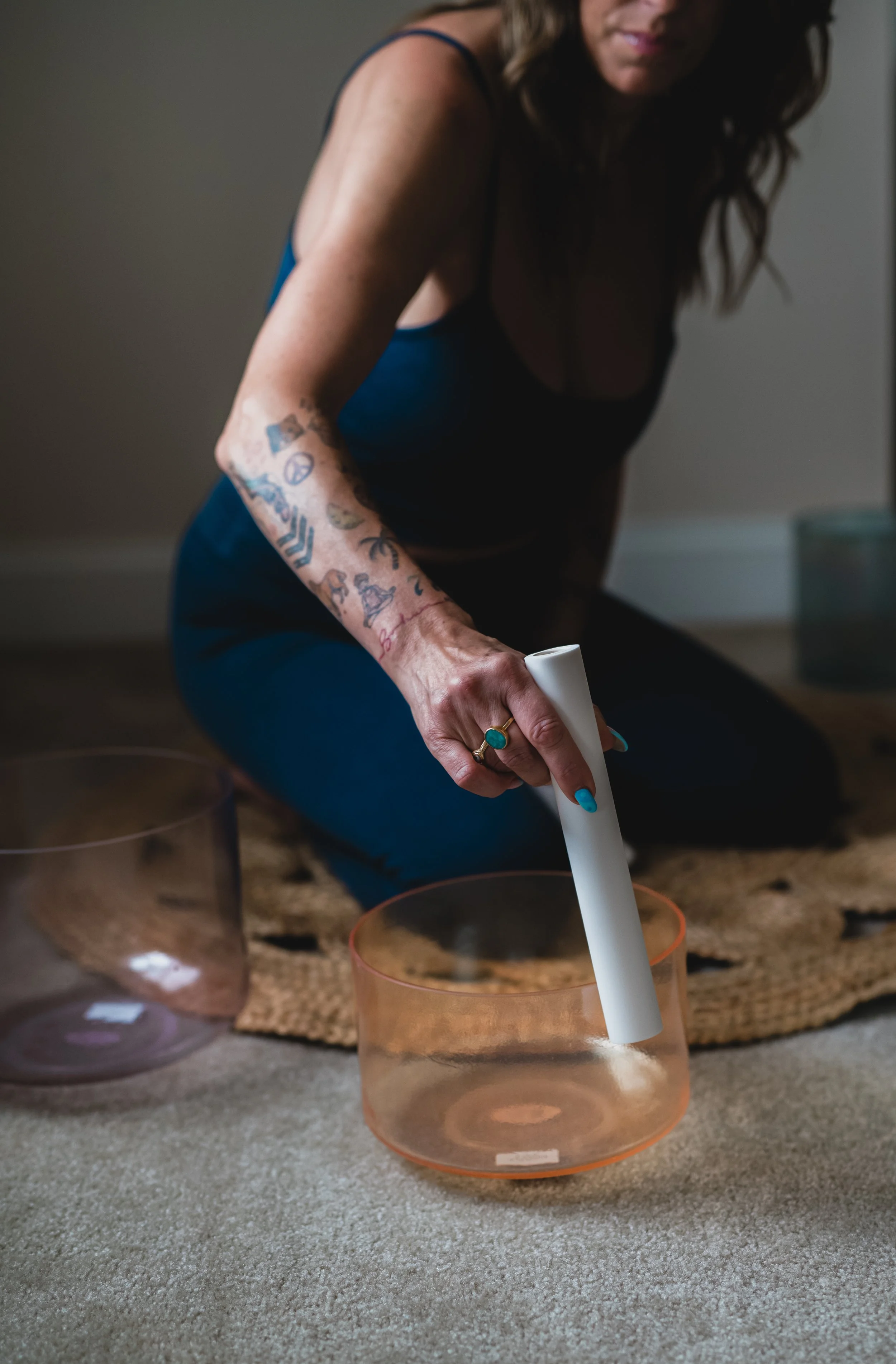 A woman with tattoos on her arm is placing a white candle into a transparent orange container on a carpeted floor. In the background, there are more similar containers and a woven mat.