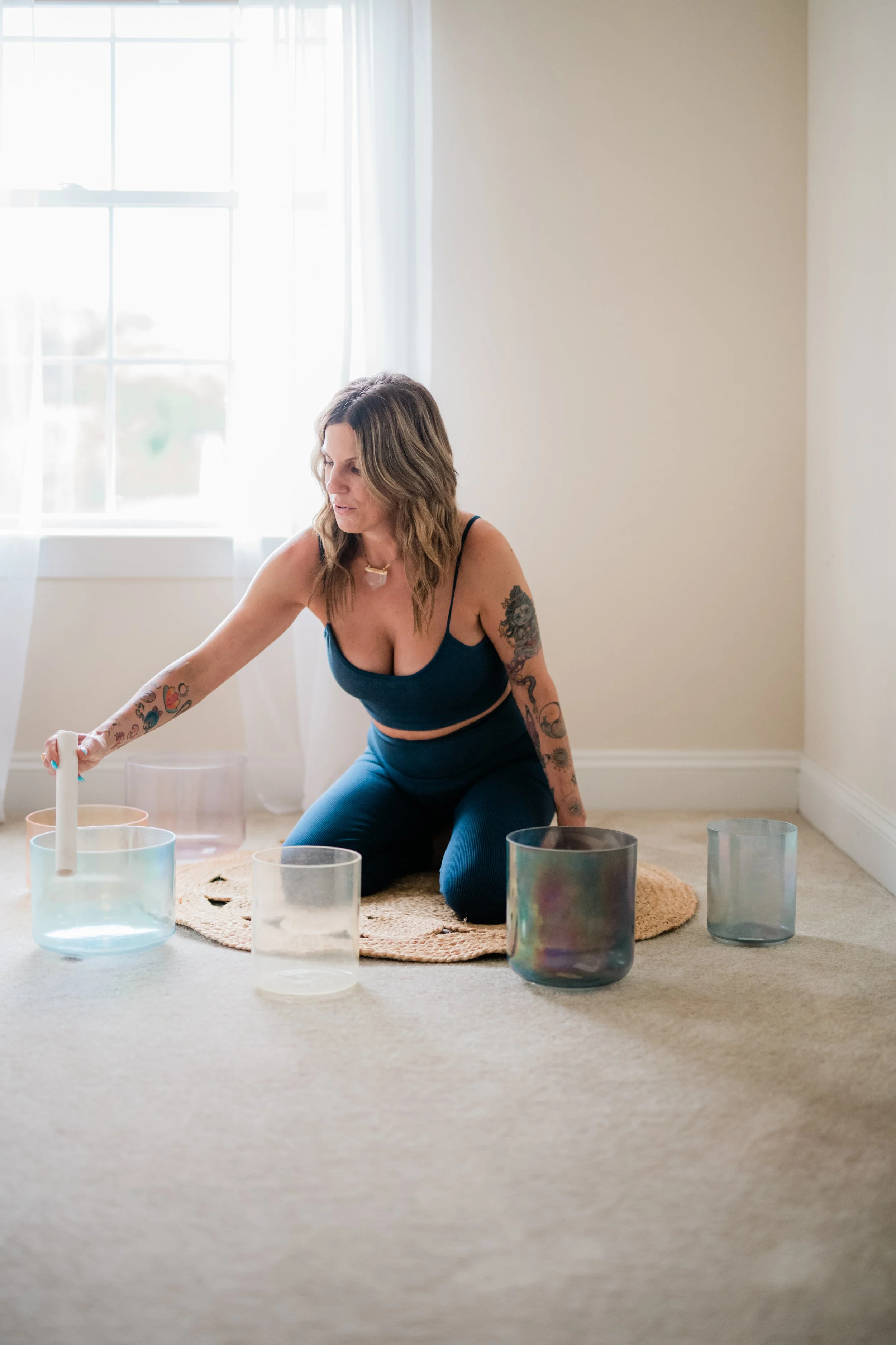 A woman with tattoos wearing a blue top and blue pants, sitting and leaning forward, surrounded by colorful glass bowls, on a beige carpet in a room with white curtains and a large window.