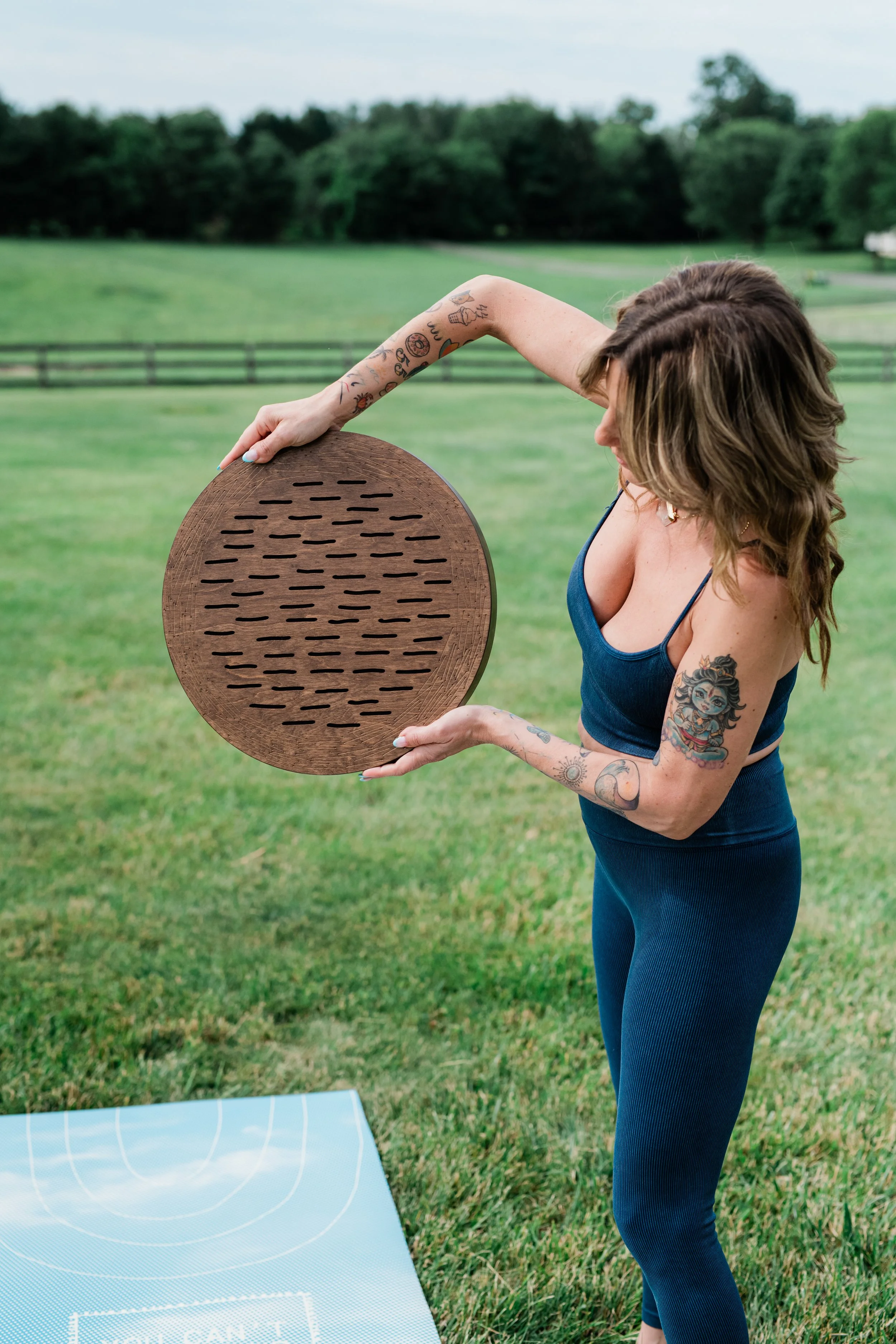 Woman practicing yoga outdoors on a grassy field holding a wooden sound bowl with both hands, wearing a blue tank top and dark blue leggings, with tattoos on her arms and a tattooed shoulder, and a yoga mat in front of her.