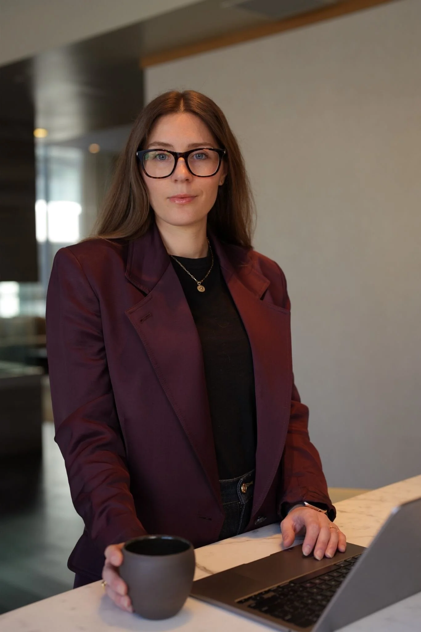 A woman with long brown hair, glasses, a burgundy blazer, and gold jewelry holding a coffee cup in one hand and working on a laptop at a counter in a modern, well-lit space.