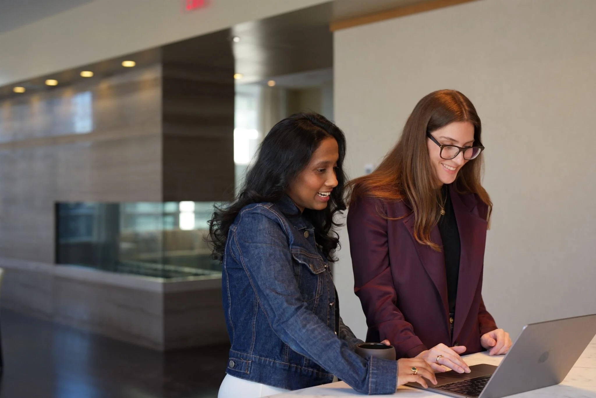 Two women are standing at a counter, looking at a laptop and smiling. One woman has long, wavy black hair and is wearing a denim jacket, while the other has long, straight brown hair and glasses, and is dressed in a maroon blazer. They are in a modern, well-lit indoor space with a metallic and concrete background.