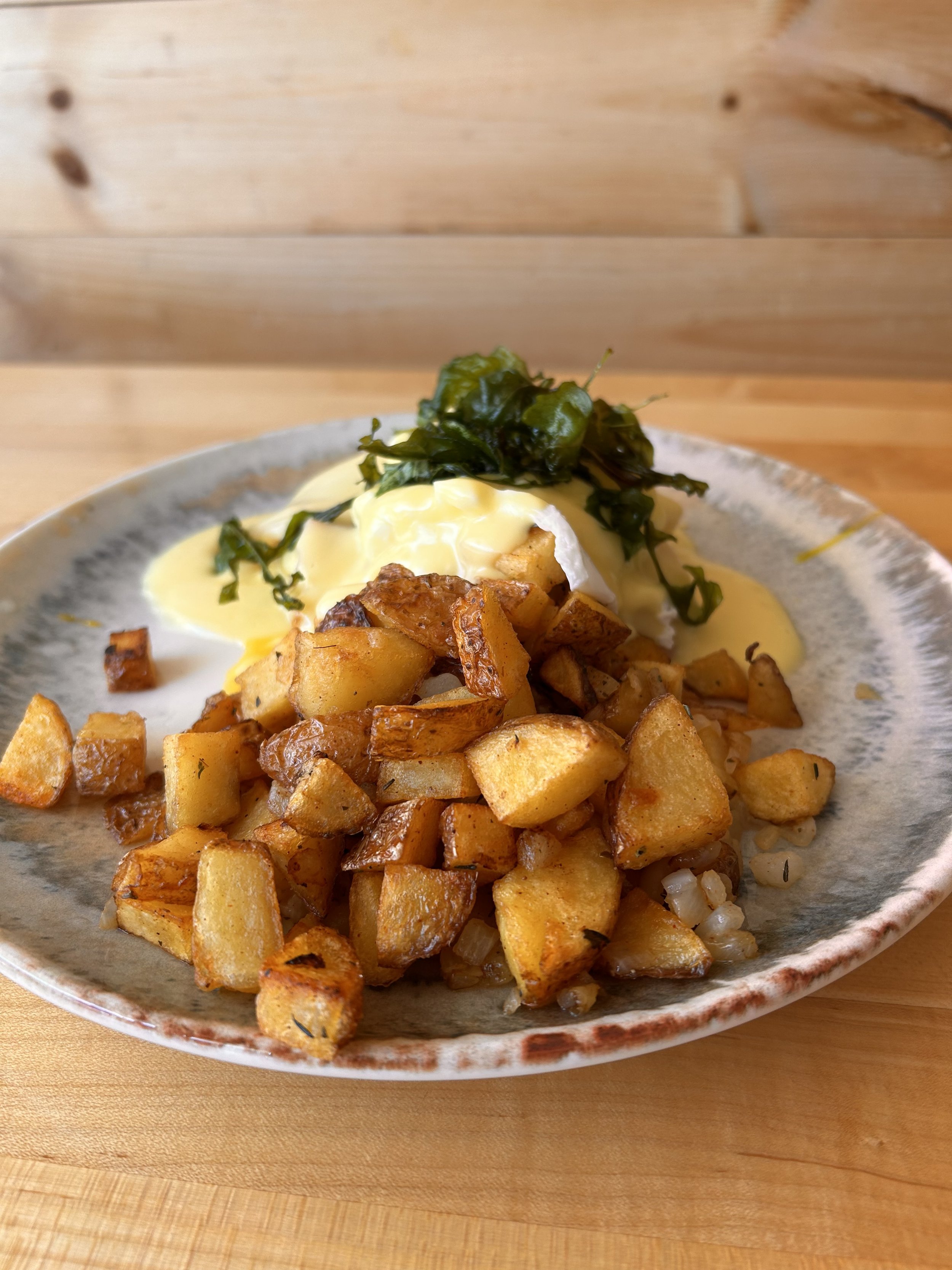 A plate of breakfast food with diced potatoes, eggs, and cooked greens, garnished with sauce, on a ceramic plate on a wooden surface.
