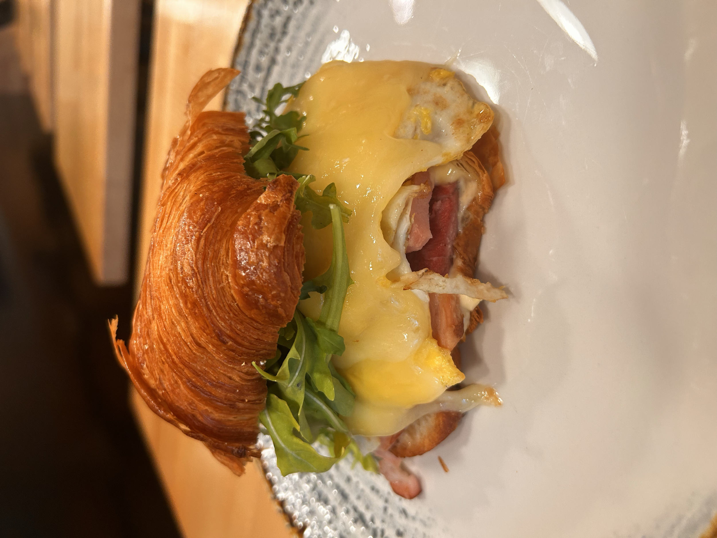 A savory breakfast dish featuring a croissant sandwich with cheese, a fried egg, bacon, and fresh greens on a white plate.