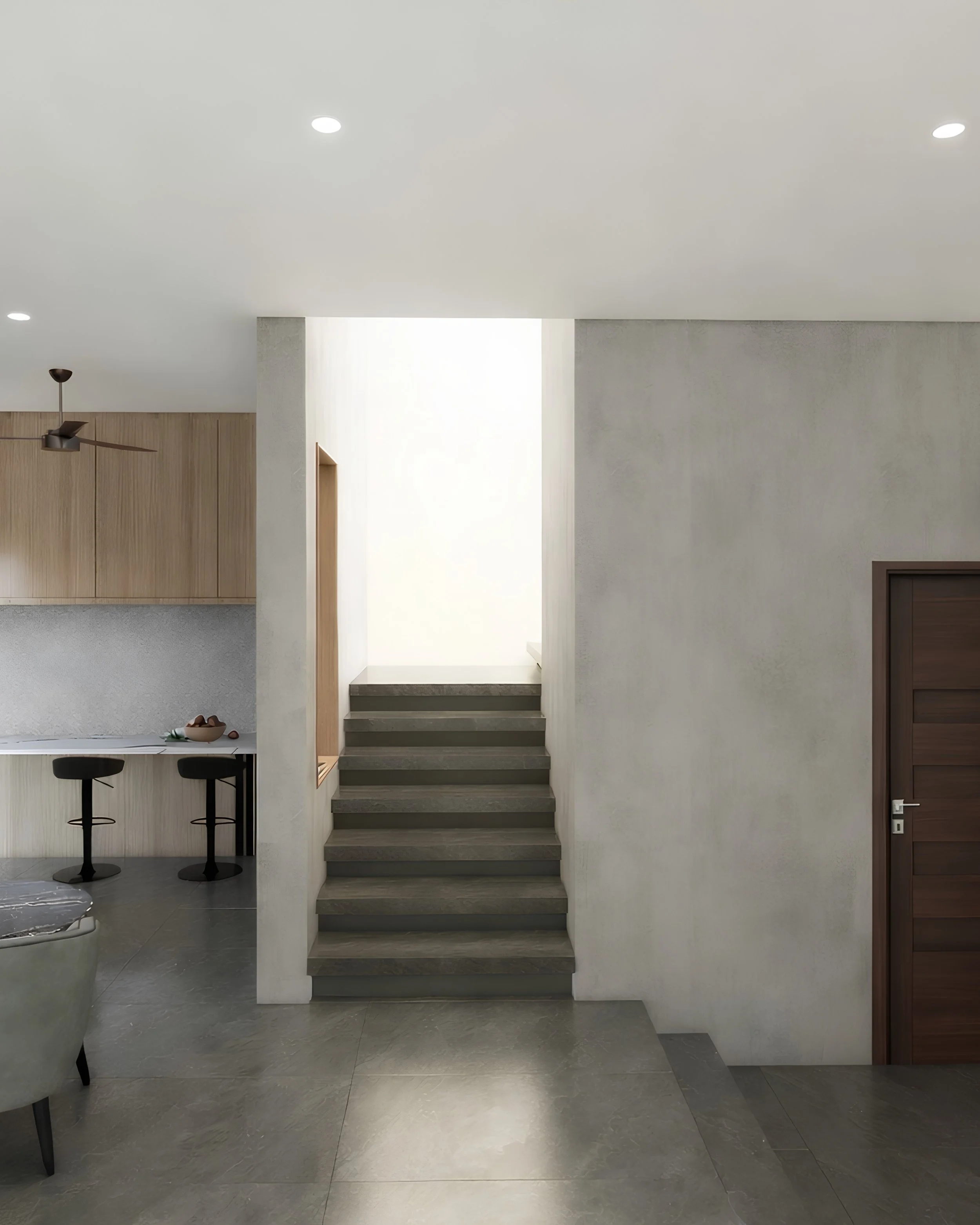 Interior view of a modern home with a staircase, gray flooring, a white wall, and a wooden door on the right. There is a built-in kitchen counter with two black bar stools with round seats and a black ceiling fan with three blades.