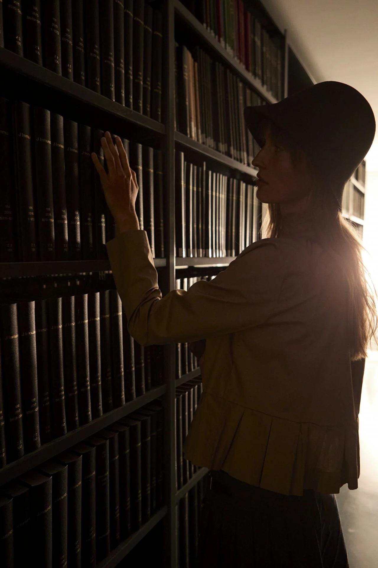 A woman with a hat browsing books or files on bookshelf in a dimly lit library or archive.