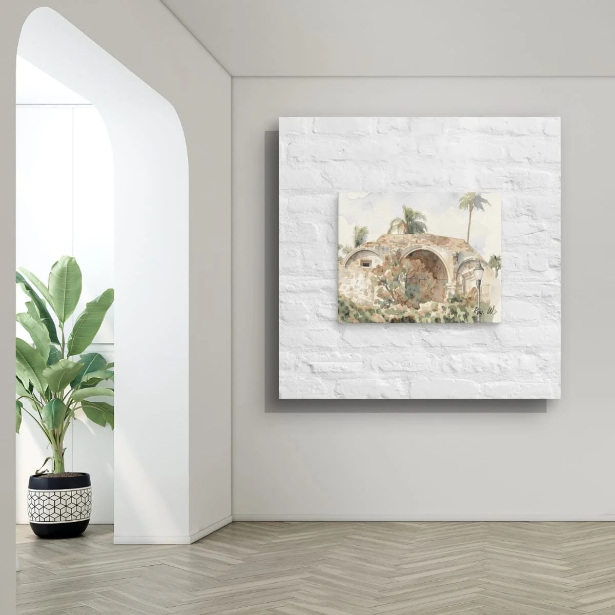 A white brick wall with a landscape watercolor painting, of San Juan Historical Church corner with a large green potted plant, and a light wood herringbone floor.