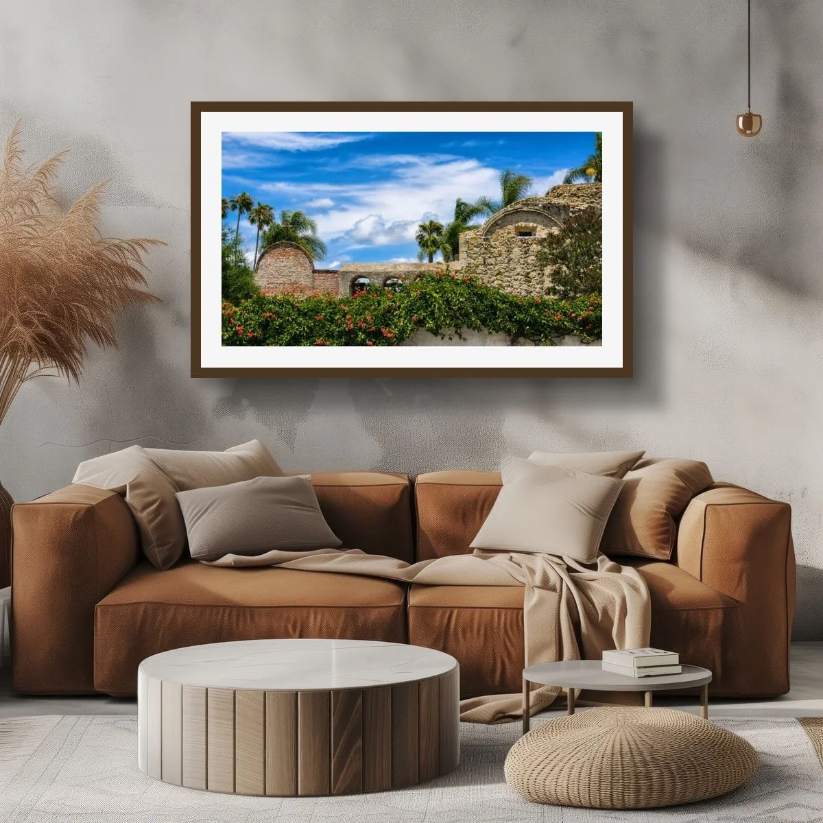 Living room with a brown sofa, beige pillows and throw blanket, round marble coffee table, small side table with books, beige pouf, large framed landscape photo of ruins and palm trees on the wall, decorative dried plants in corner.