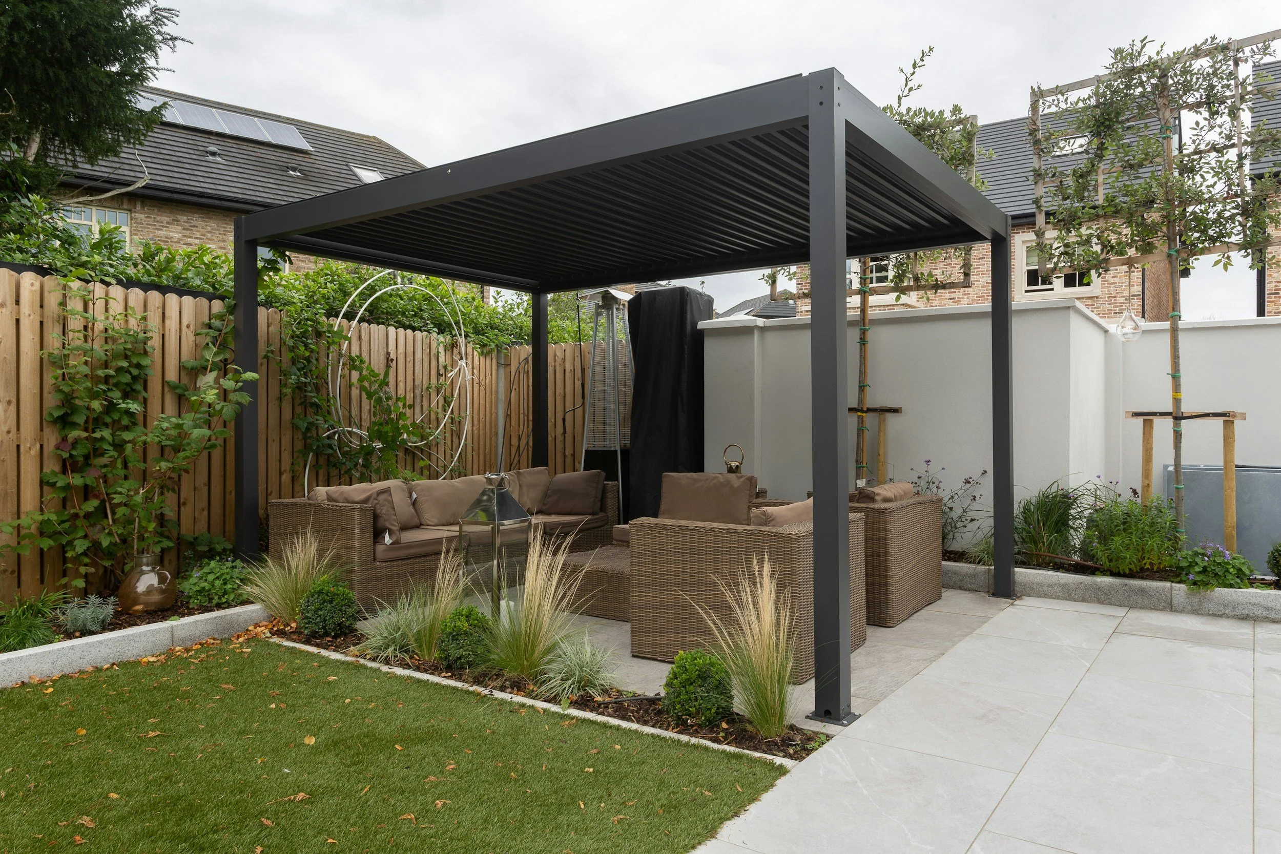 Backyard with a modern pergola, beige outdoor furniture, green plants, a wooden fence, a lawn, and neighboring brick houses.