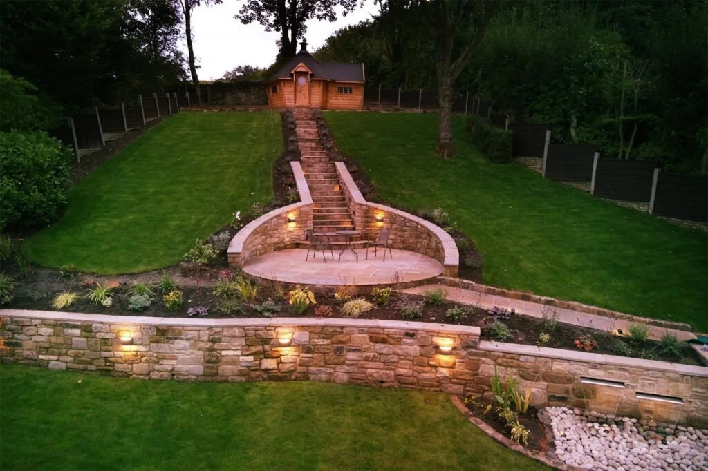 A landscaped backyard with a staircase leading up to a small wooden shed, stone walls with embedded lights, lush green grass, and surrounding trees.