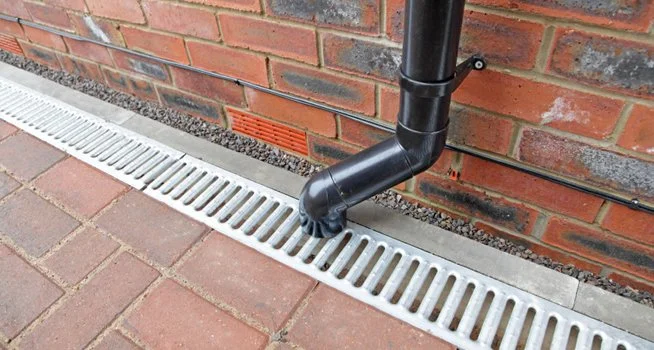Black drainpipe attached to a red brick wall, directing water into a metal drainage grate on a sidewalk.