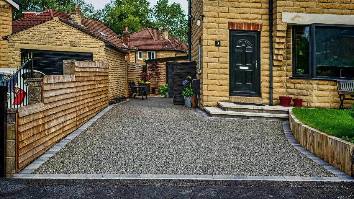 A residential driveway with a pebble finish, bordered by brick and wooden fencing, leading to a yellow brick house with a black front door and potted plants, a bench, and a grass lawn with a curved wooden edge.