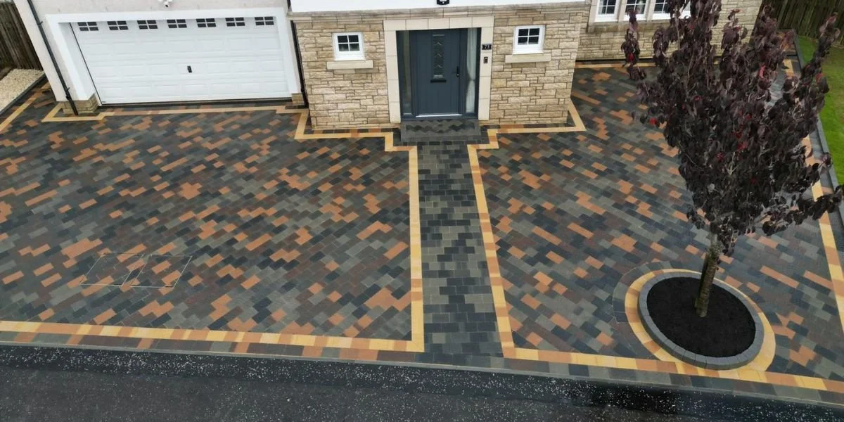 A newly paved driveway with multicolored brick pavers, a central walkway leading to a front door, a tree planted in a round bed, and the front of a house with stone and siding exterior.