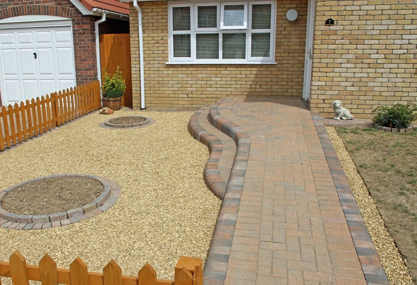 A front yard with a curved brick walkway, two circular planting beds with bricks lining the edges, a small dog statue, a potted plant, and a wooden fence on the left. The house has beige brick walls, a window, and a garage door.