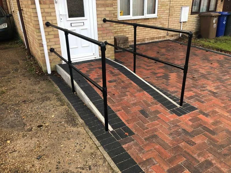 A wheelchair accessible ramp with black metal railings leading to a white door on a brick patio in front of a brick house.