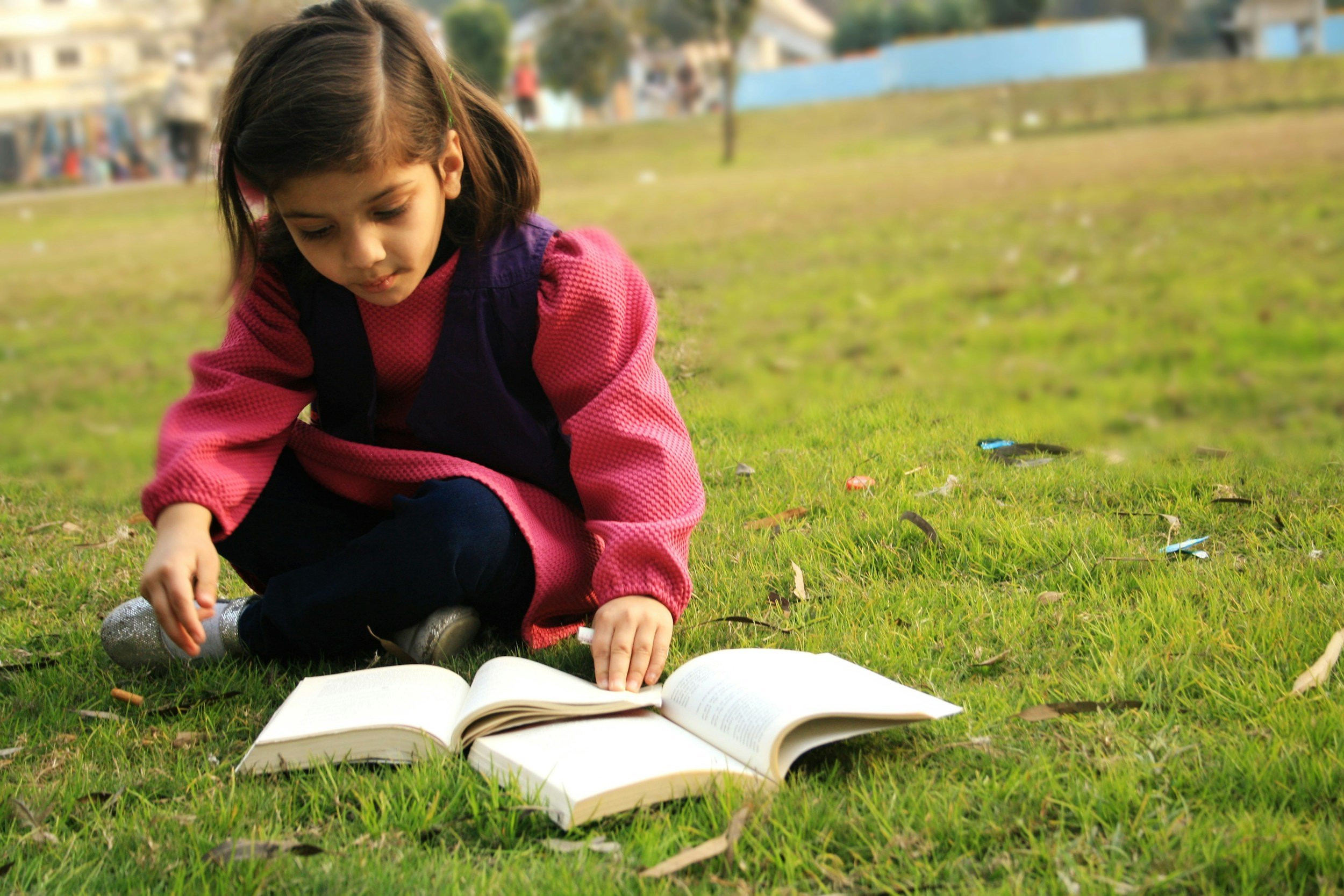 Young girl sitting on grass in a park, reading a book with colorful papers scattered around her.