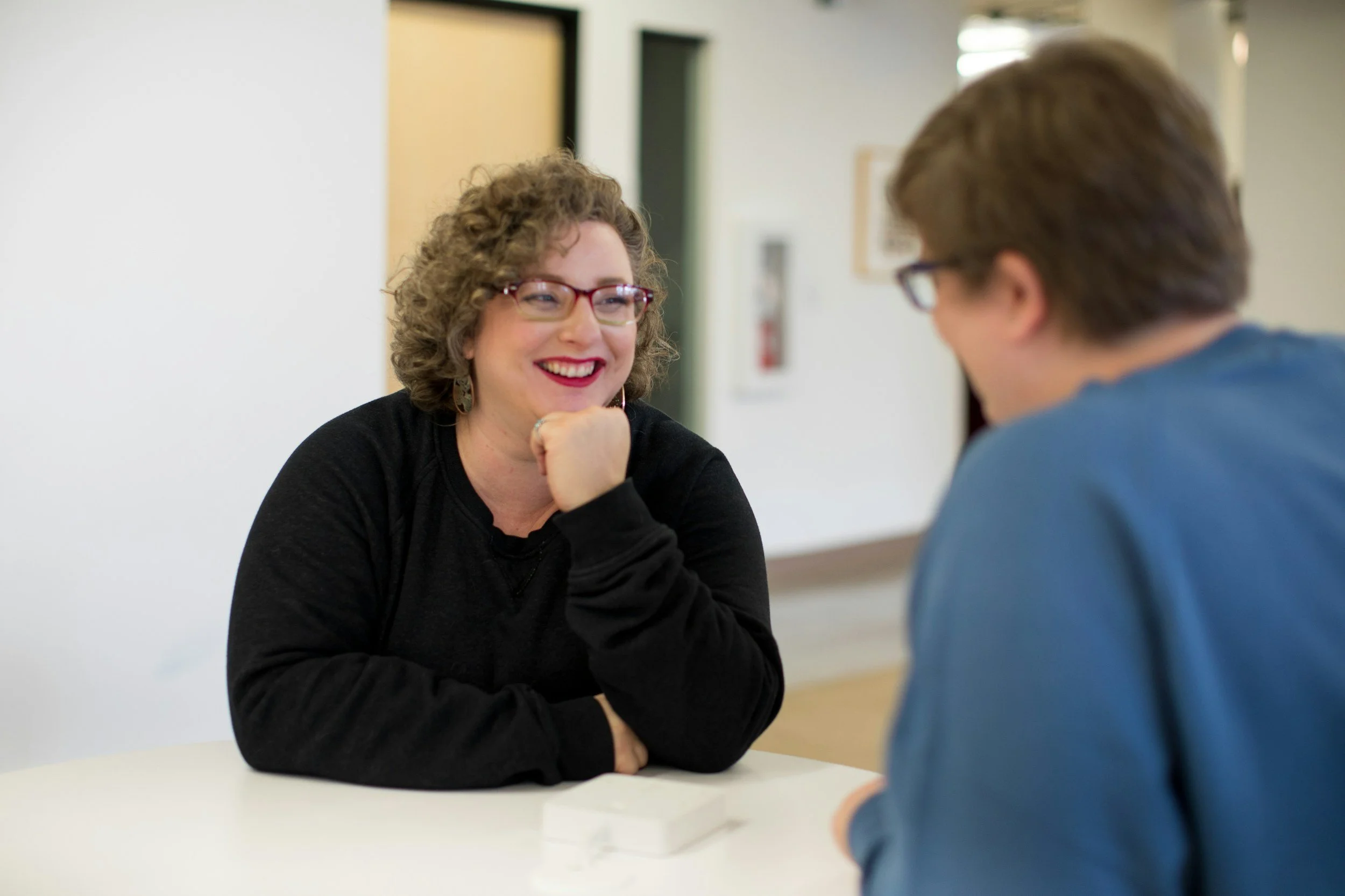 A speech and language pathologist with curly hair and glasses smiling and engaging in conversation therapy with a man in a blue shirt in an indoor setting.