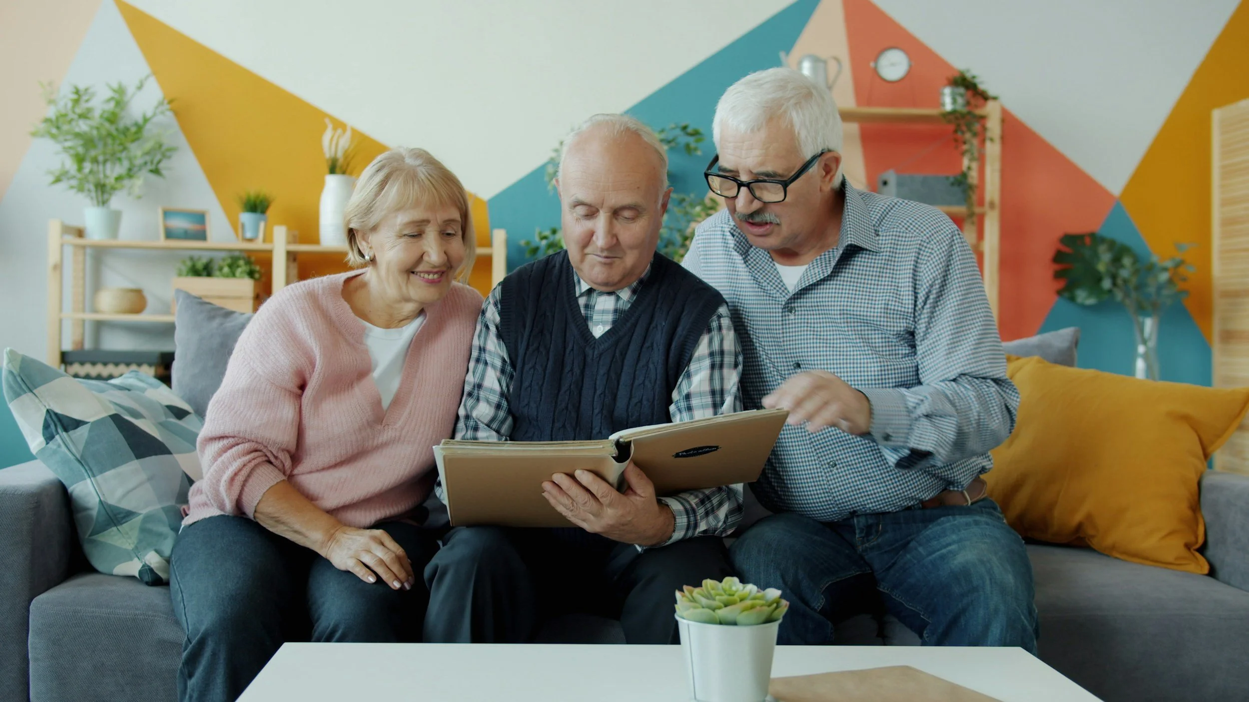 Three elderly people sitting on a couch looking at a photo album together in a brightly decorated living room with colorful geometric wall art, completing speech therapy activities.