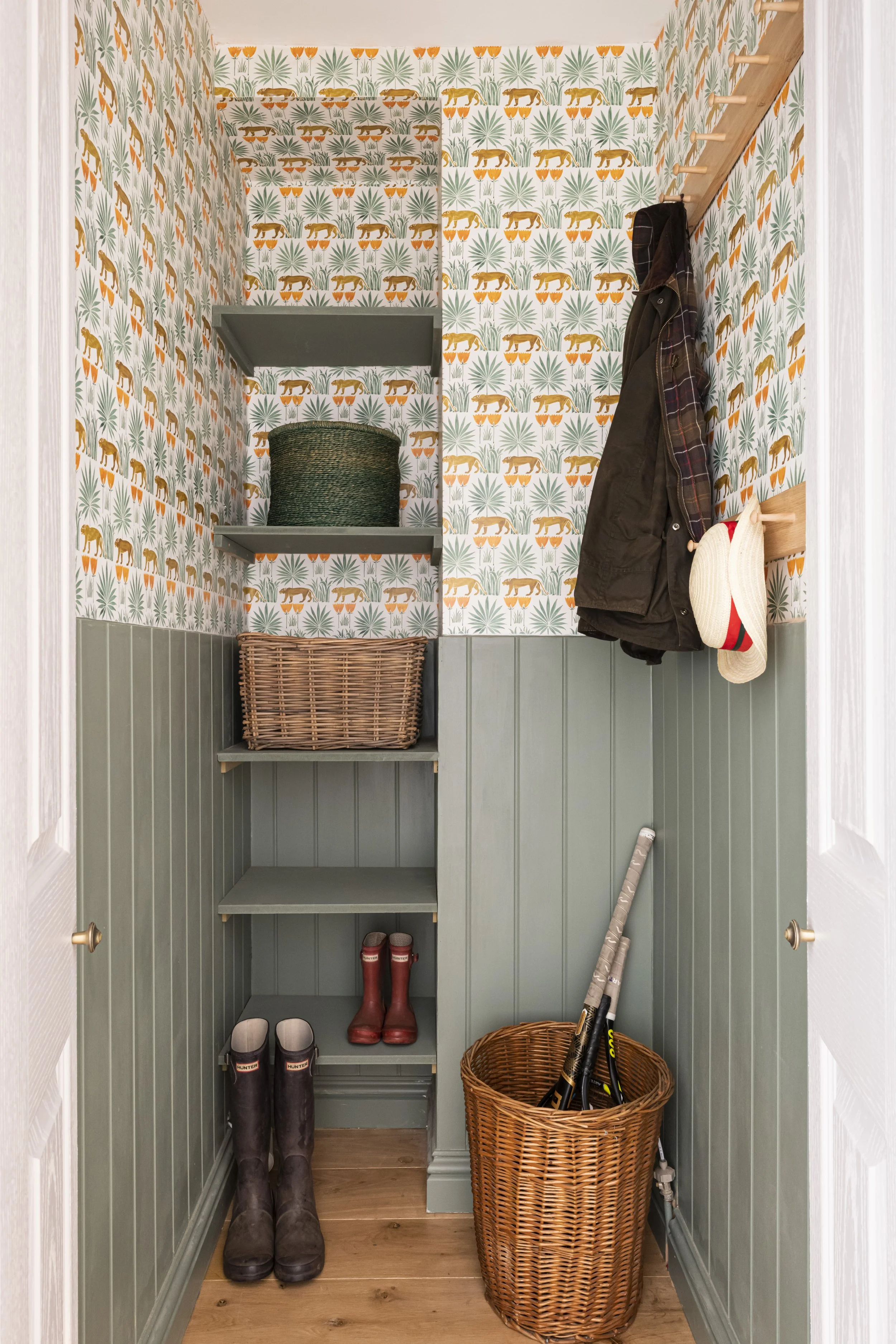 A small entryway closet with green beadboard walls, patterned wallpaper with animal and plant motifs, gray shelves holding a woven basket and black container, hanging coats and hats, and shoes and boots on the floor.