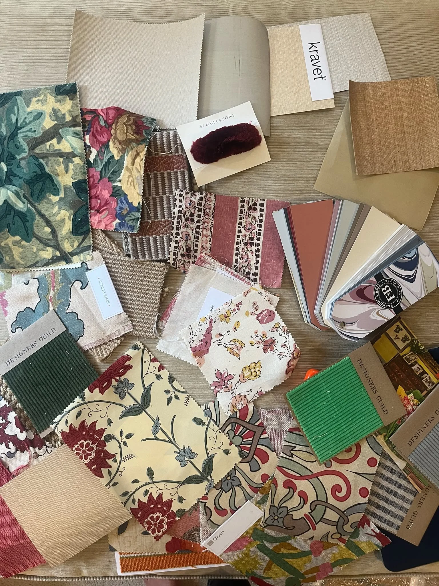 A beige wall with various items attached, including dried flowers, paper notes, fabric swatches, a wooden frame, and a pink vase with purple dried branches. A desk at the bottom holds a clipboard, pen, and small decorative objects.