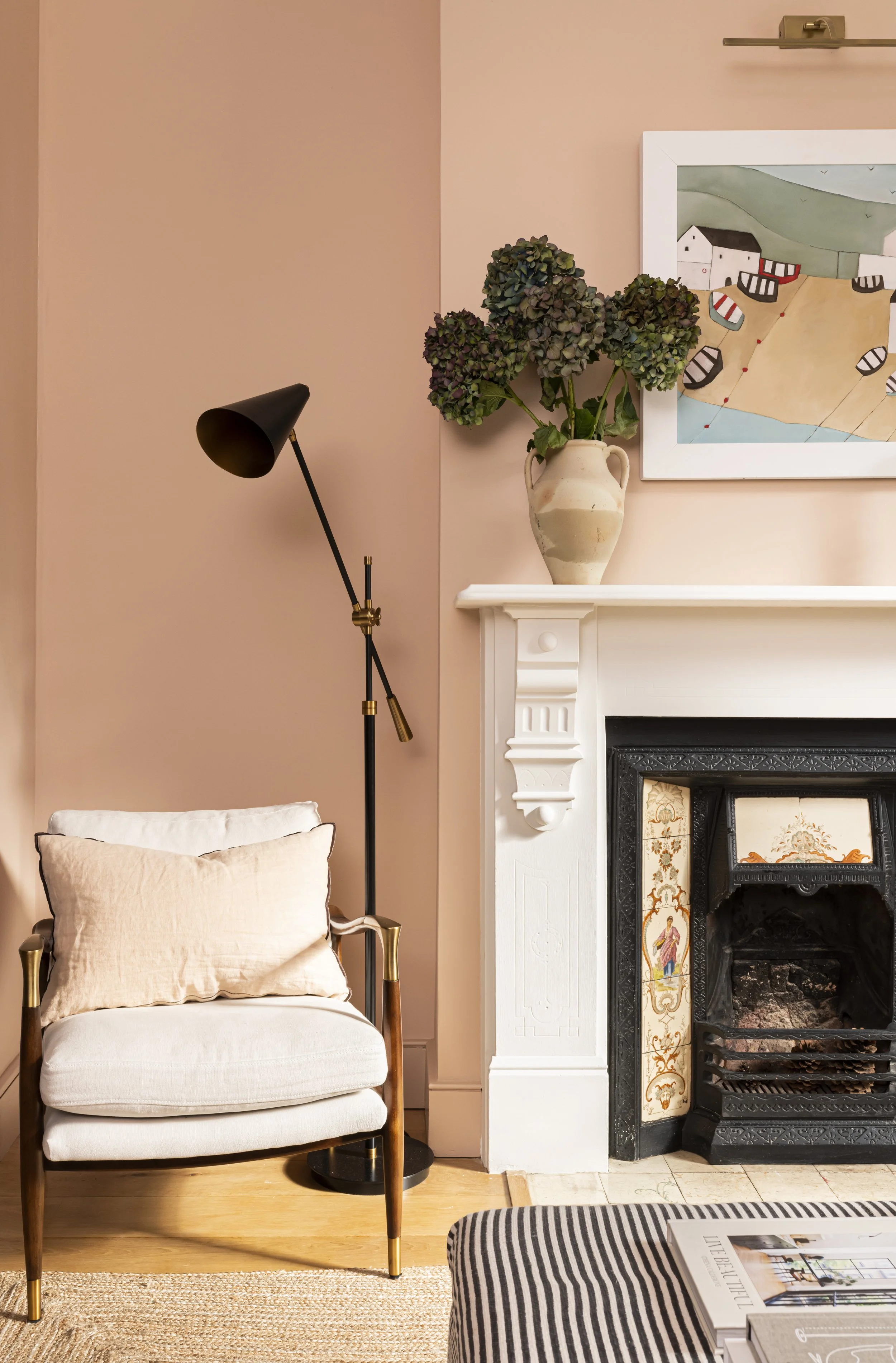 Living room with a beige armchair, a black and gold floor lamp, a painting of a seaside village, a mantel with a vase of purple hydrangeas, a fireplace with decorative tiles, and a striped ottoman with magazines.