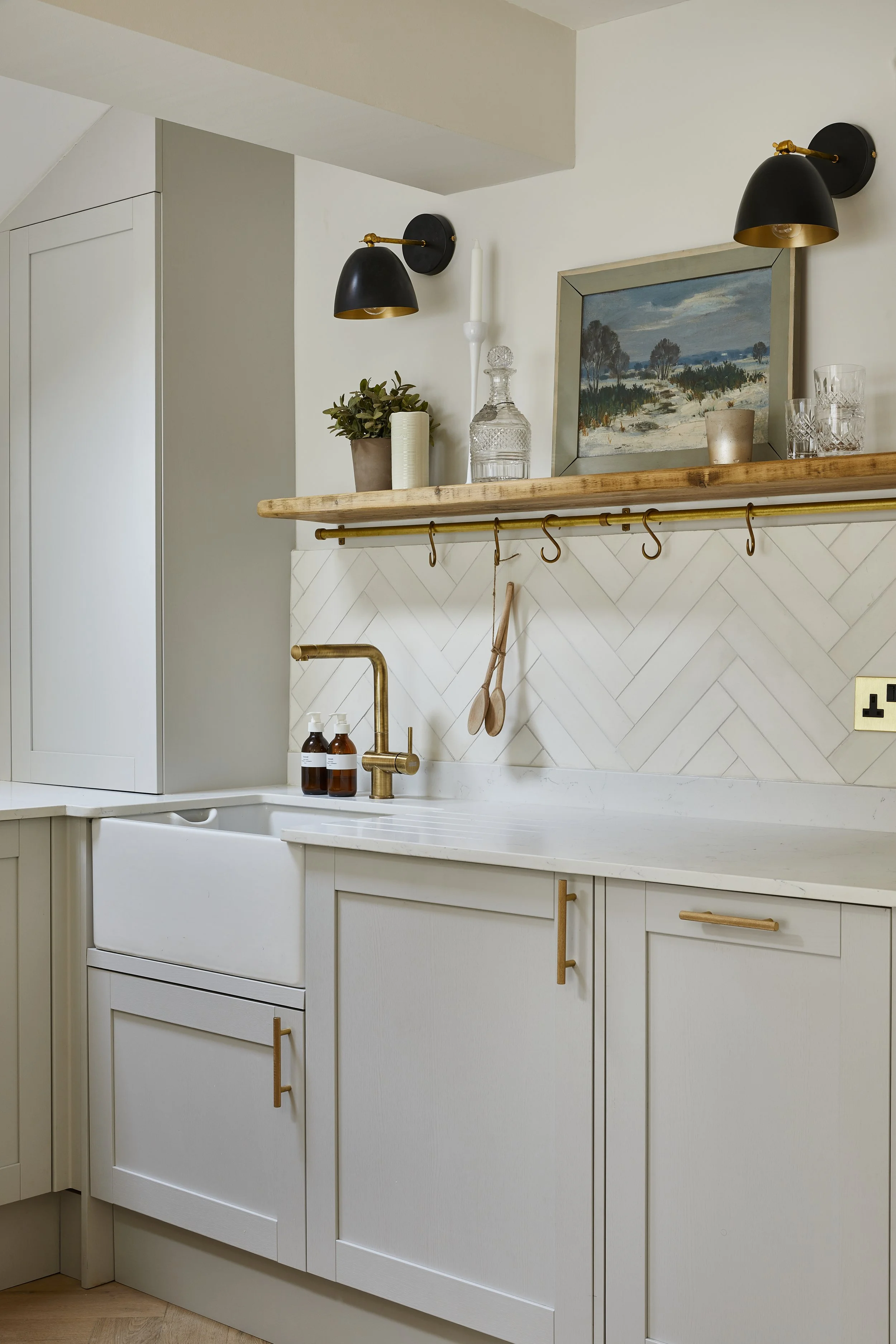 A kitchen with white cabinetry, gold cabinet handles, a gold faucet, and a tiled backsplash. Open shelving displays wine glasses, a painting of a landscape, and decorative items. Black wall sconces with gold accents are above the shelves.
