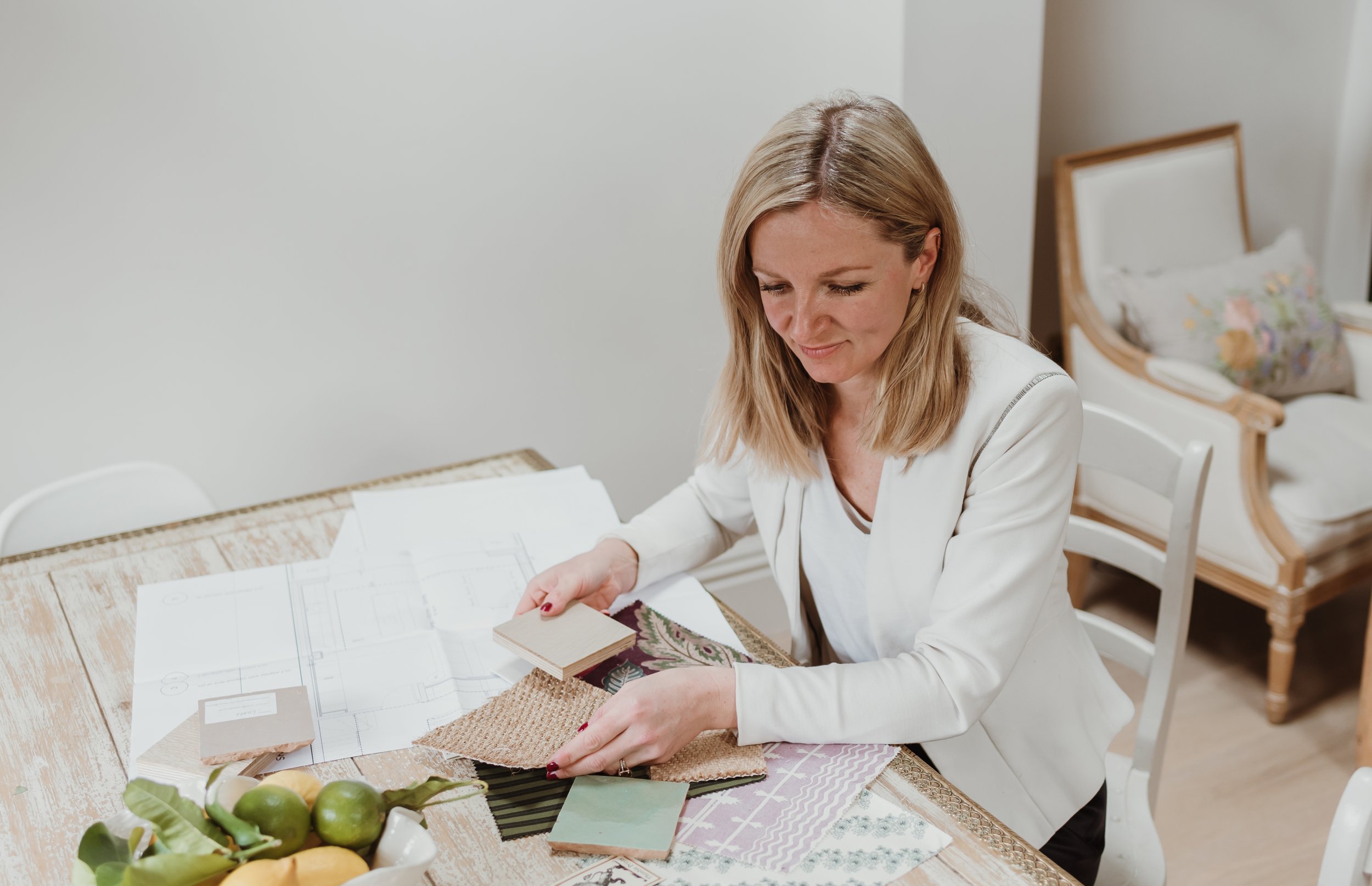 A woman with shoulder-length blonde hair looks at fabric samples at a table. There are sketches, fabric swatches, and color samples on the table, with a bowl of lemons and limes in the foreground. A bed with floral pillows is visible in the background.