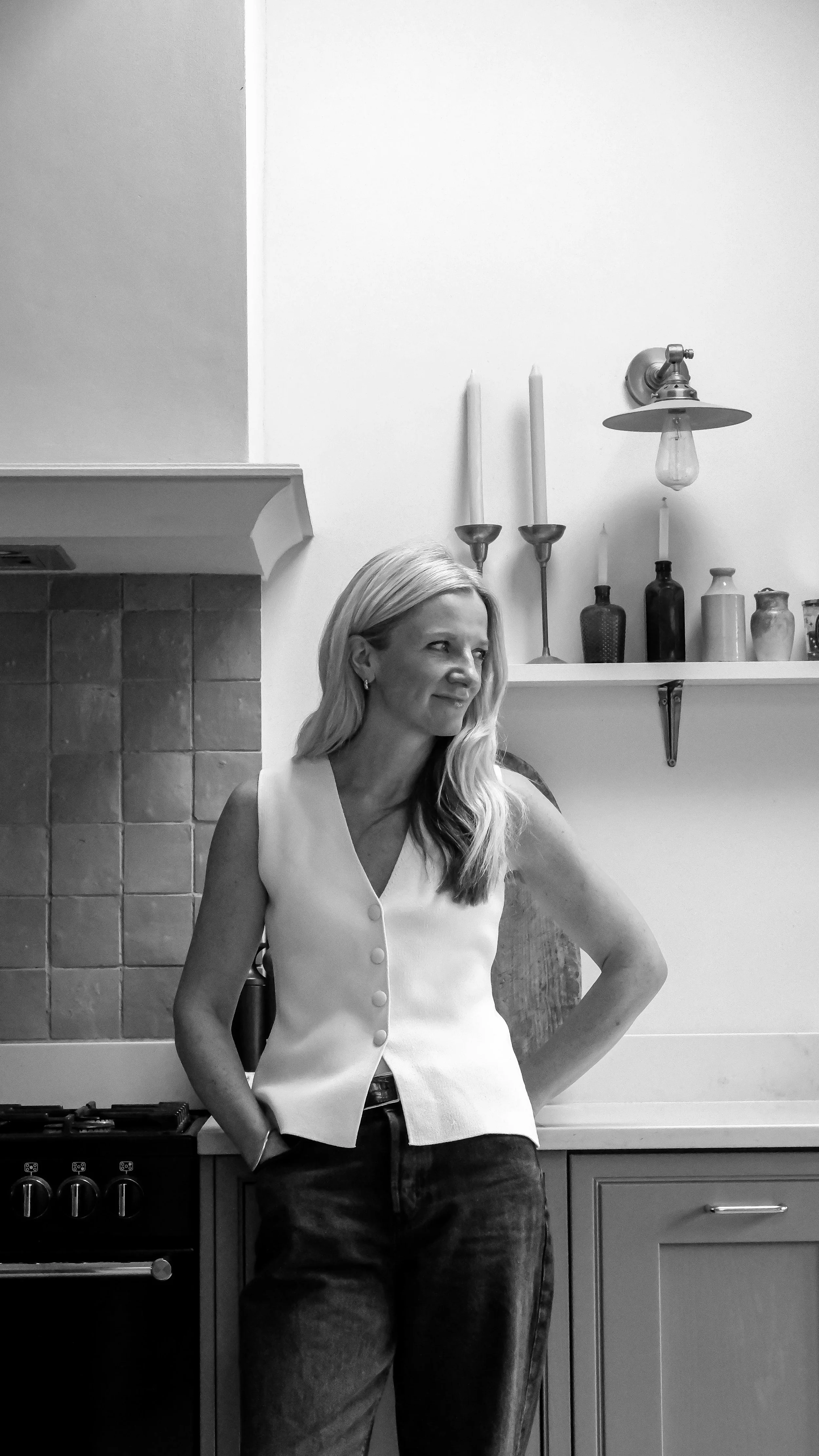 A woman with long blonde hair wearing a sleeveless vest and jeans, standing in a modern kitchen with a stove, tiled backsplash, and decorative bottles on a shelf.