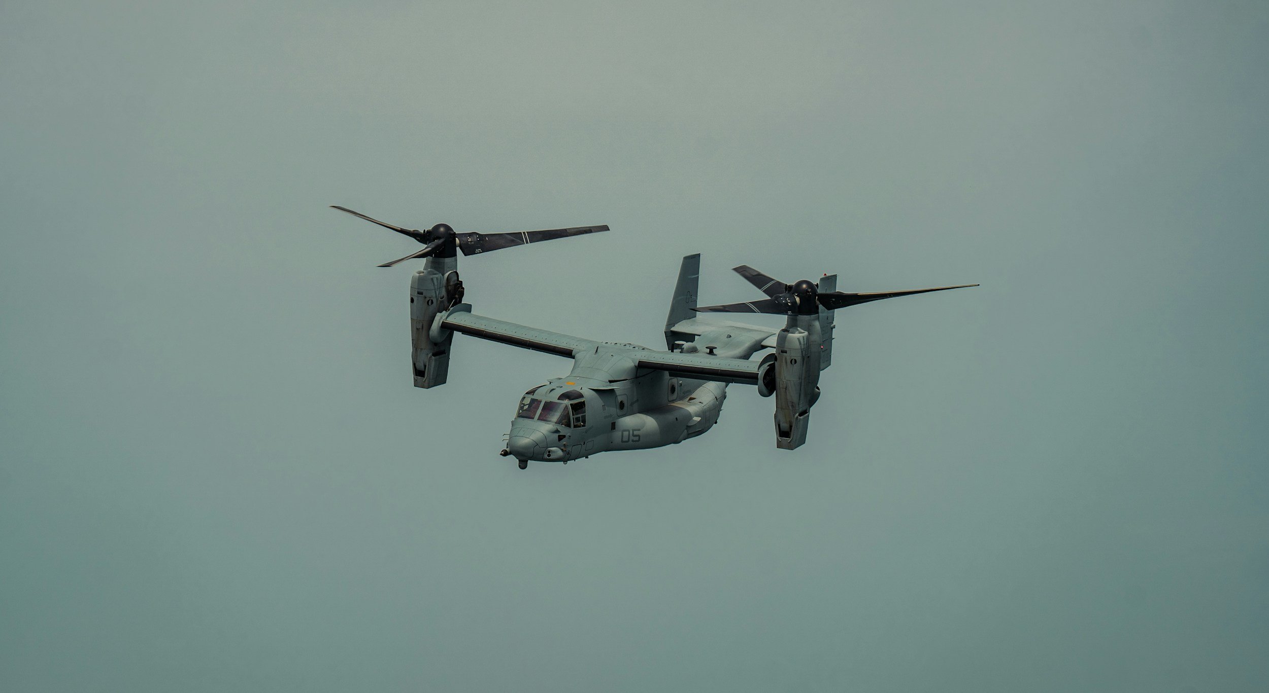 A military-style helicopter, likely a V-22 Osprey, flying in the sky with its rotors in motion.