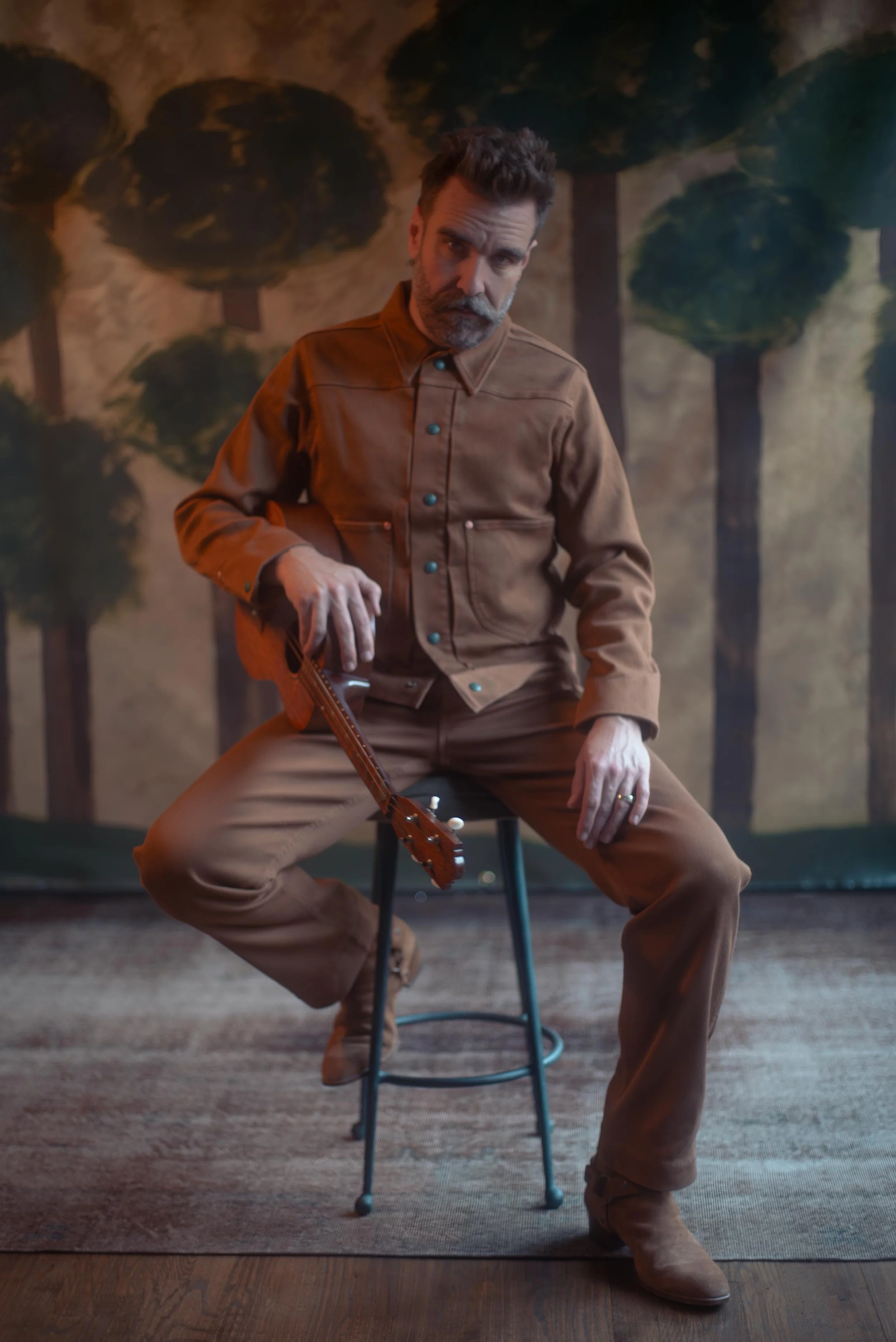 Scott Terry wearing a tan corduroy jacket and pants, poses with a rifle in front of a painted background of trees and a cloudy sky.