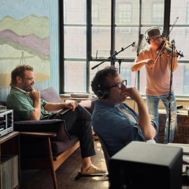 Three men in a studio, one sitting on a sofa with a drink, another wearing headphones and sitting at a desk, and the third playing a violin near a window.