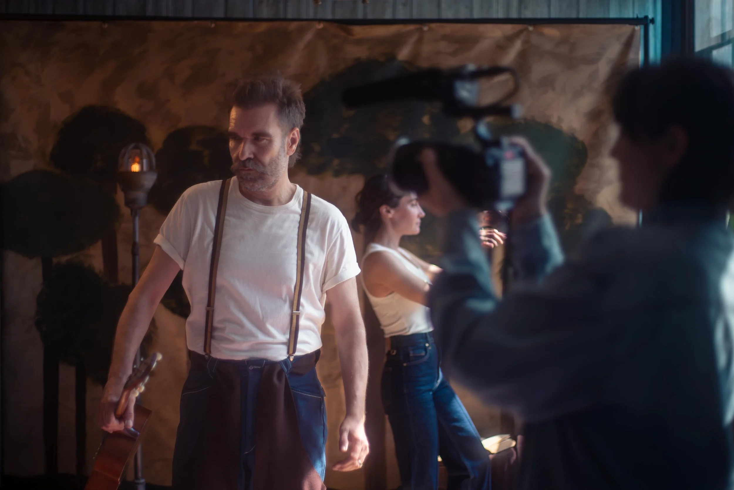 Scott Terry wearing a white t-shirt with suspenders, holding a ukelele, standing in a dark room with other people. The background shows a textured wall and natural light coming through a window.