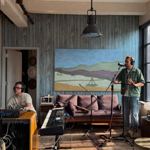 Scott Terry & Will Hoge in a room with musical equipment, Will is sitting at a control board and Scott is standing and singing into a microphone with headphones on. The room has a painting of a landscape and a window allowing natural light.