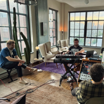 Creating the album - the crew in a sunlit urban living room with large windows, a cactus, and musical equipment, including a keyboard and sound mixer.