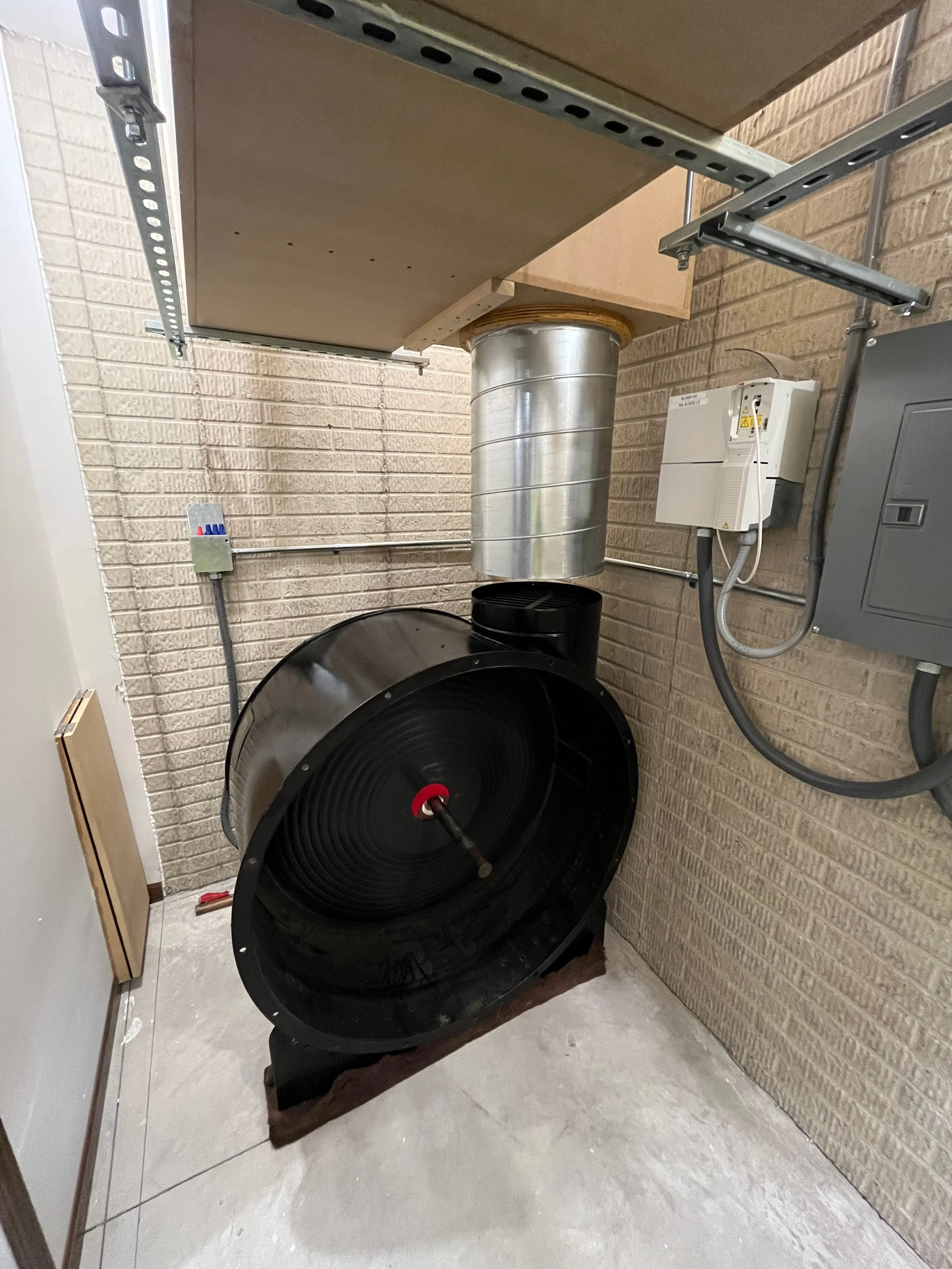 A pipe organ static wind-line connected to a large black fan, mounted in a corner of a room with brick walls, electrical boxes, and tools on the floor.
