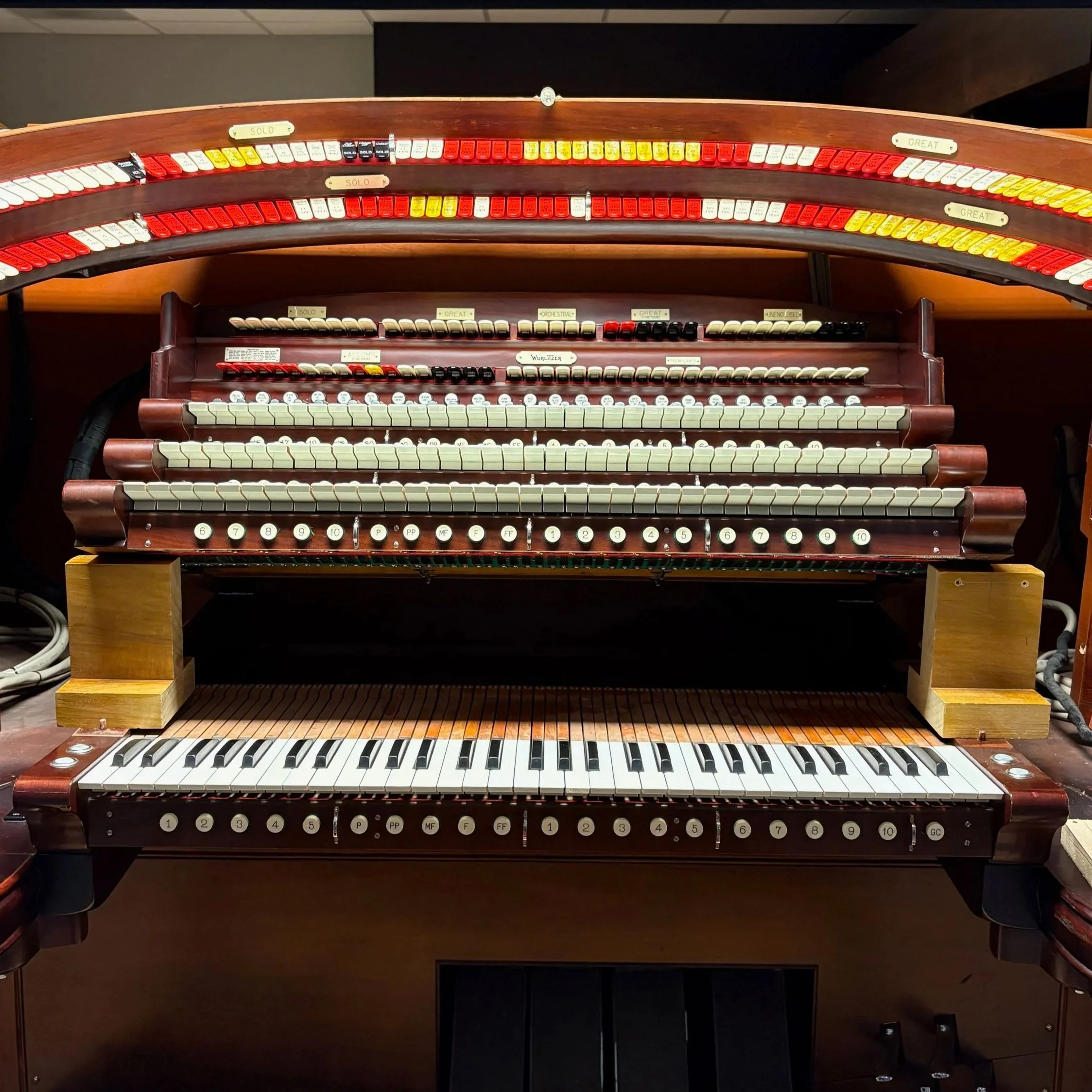 A vintage theatre organ with multiple manuals, stops, and pedals, made of dark polished wood with white, black, red, and yellow keys and stops, hinged open for mainenance.