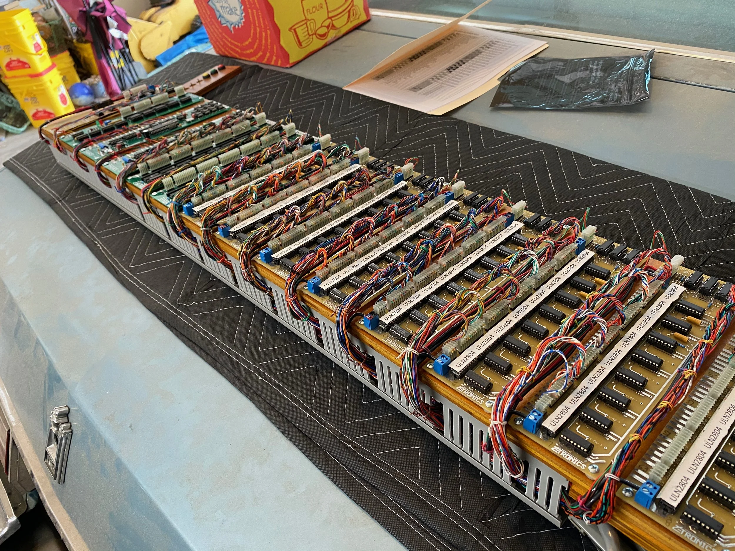 A large electronic circuit board with multiple interconnected components, wires, and chips, set on a black anti-static mat on a workbench.