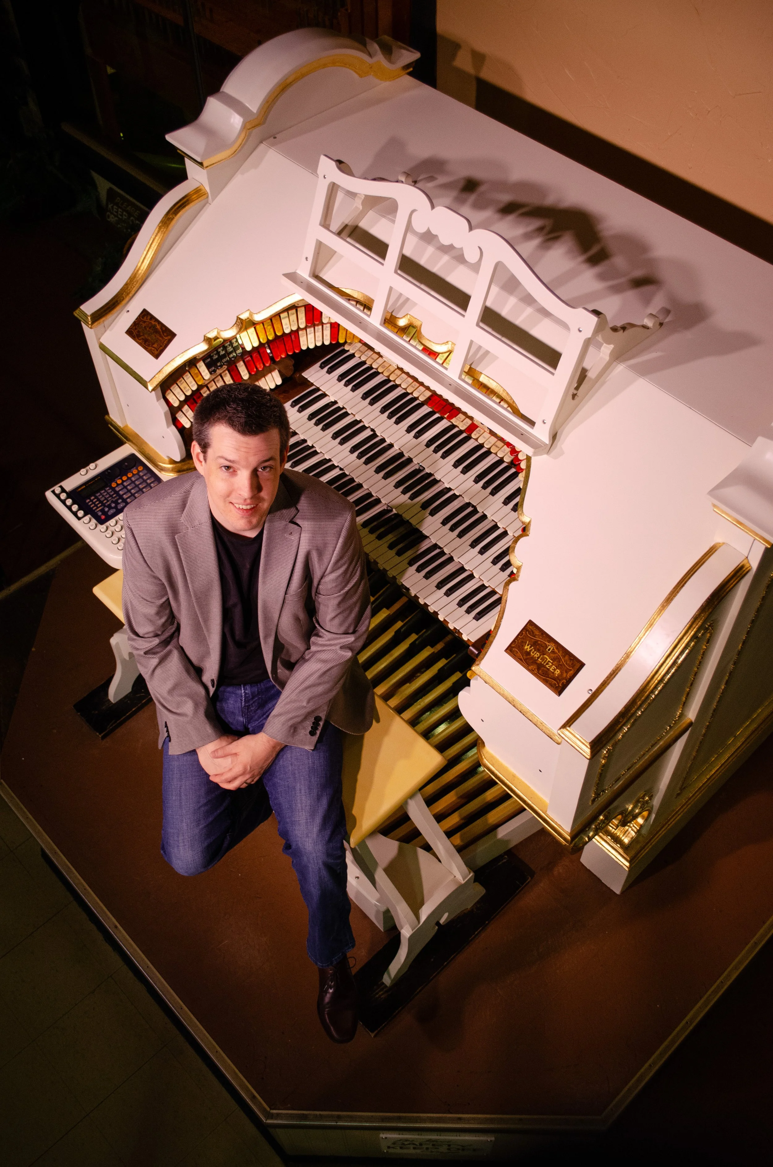 A man sitting on a bench in front of a white ornate theatre pipe organ with gold accents, inside a room with wood-paneled walls.