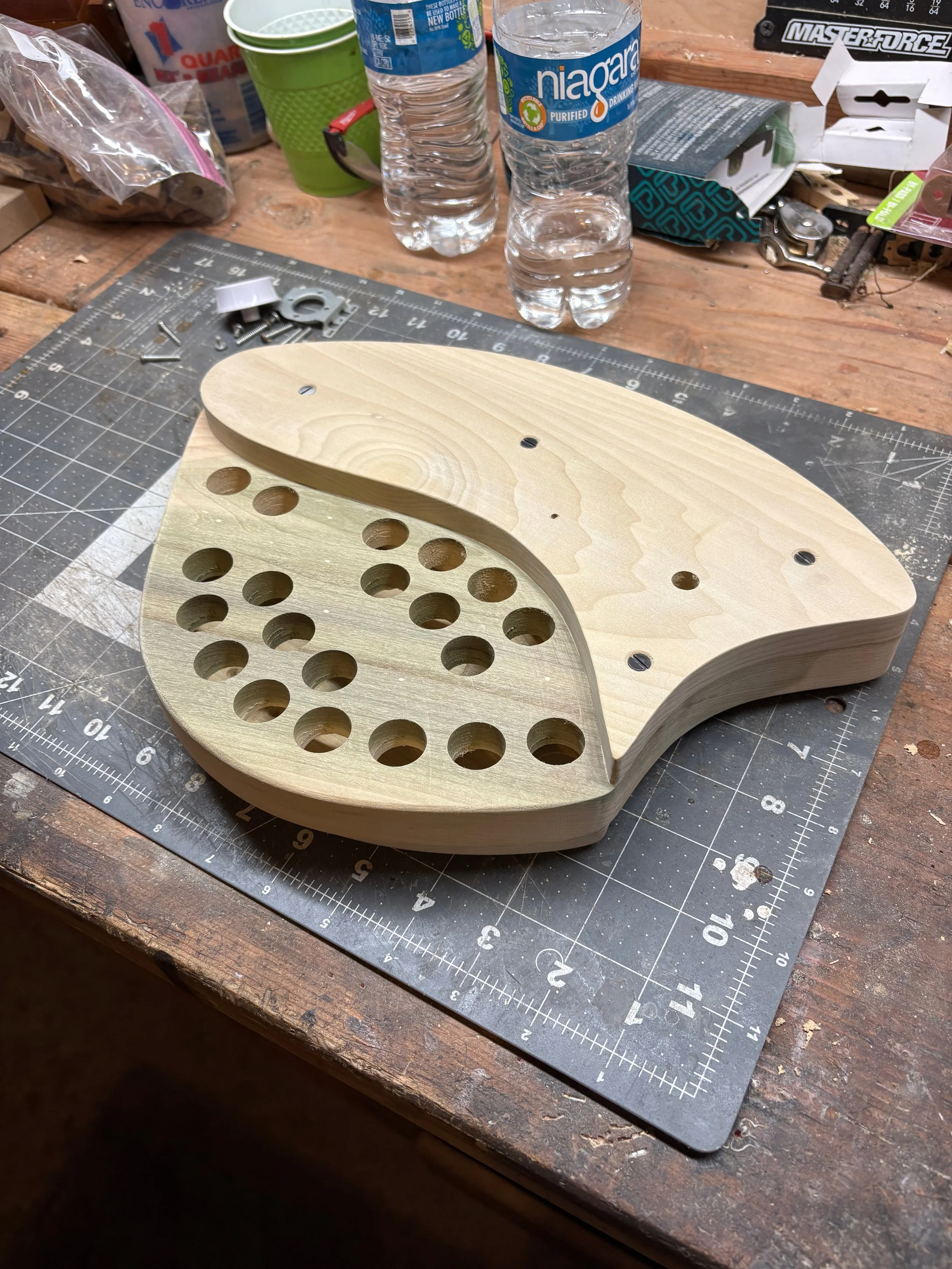 Wooden organ control panel with multiple holes, positioned on a workbench surrounded by tools, screws, and water bottles.