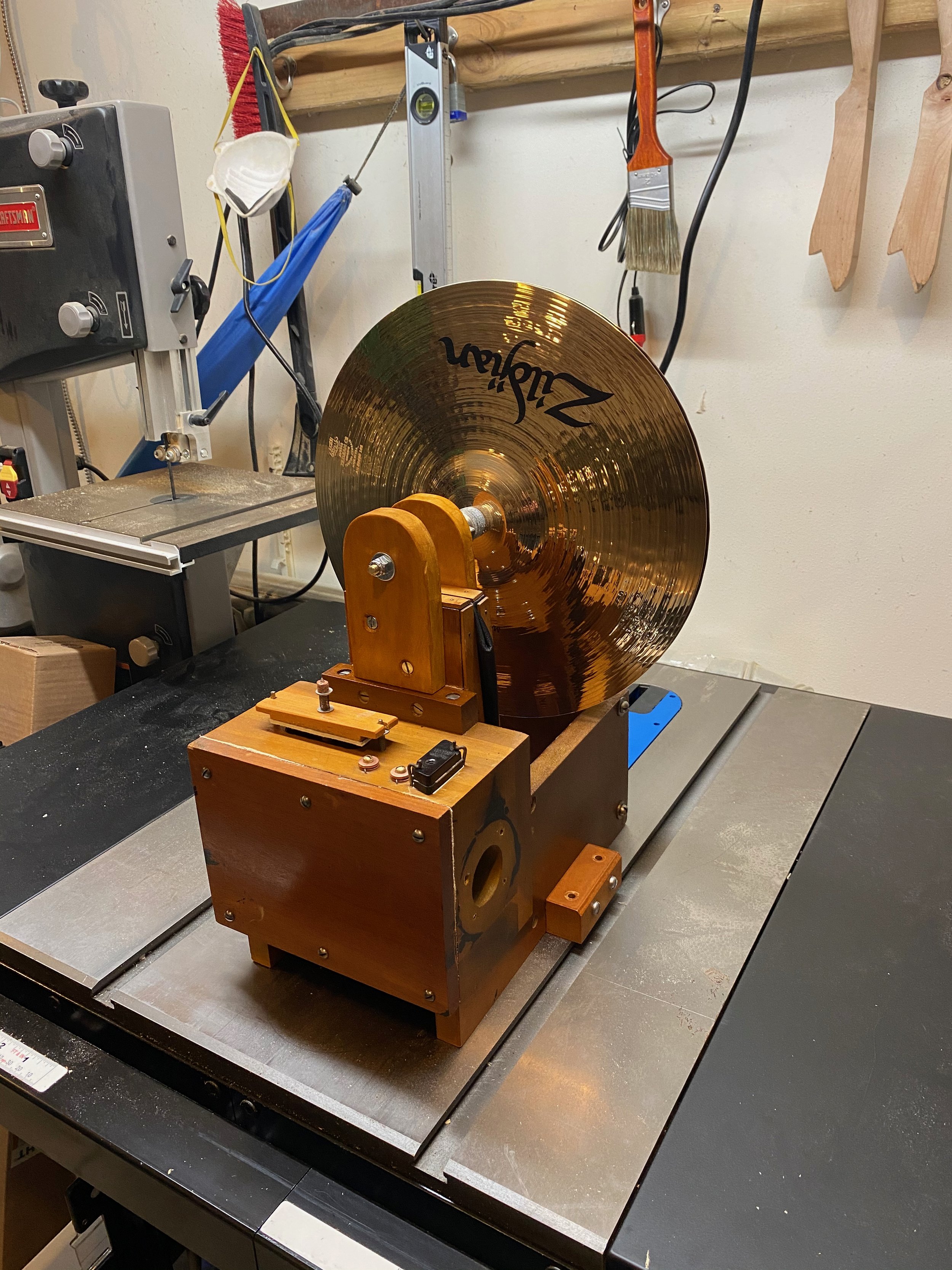 A gold-colored cymbal mounted in a workshop. The cymbal has the brand name 'Zildjian' printed on it. The background shows various tools hanging on a wall, including a paintbrush, a level, and two wooden stirrups, along with a covered wire and other workshop equipment.