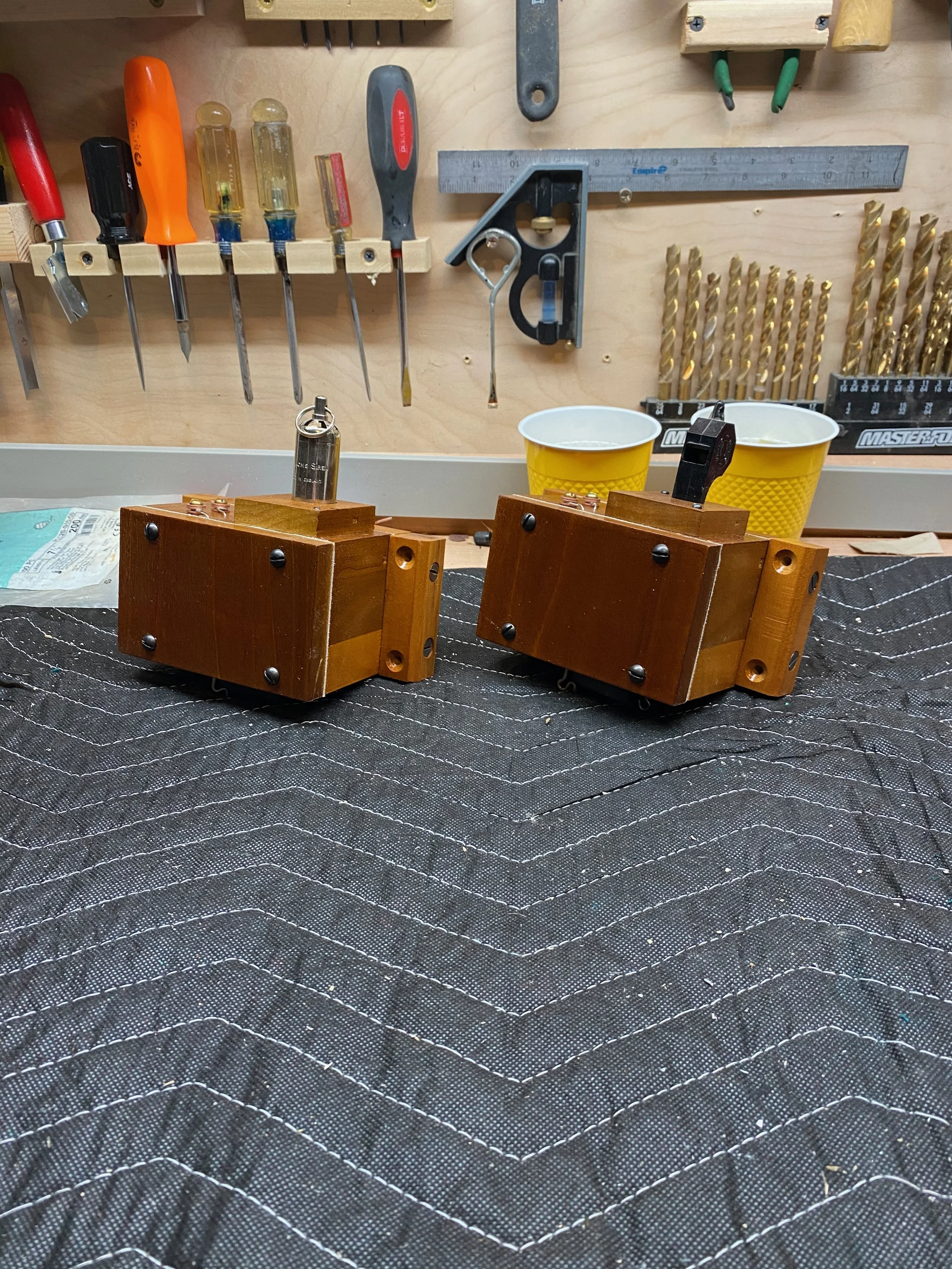 A woodworking workbench with various hand tools on a pegboard, two wooden blocks with metal fixtures, a metal square, a ruler, two yellow cups, and a black tool mat.