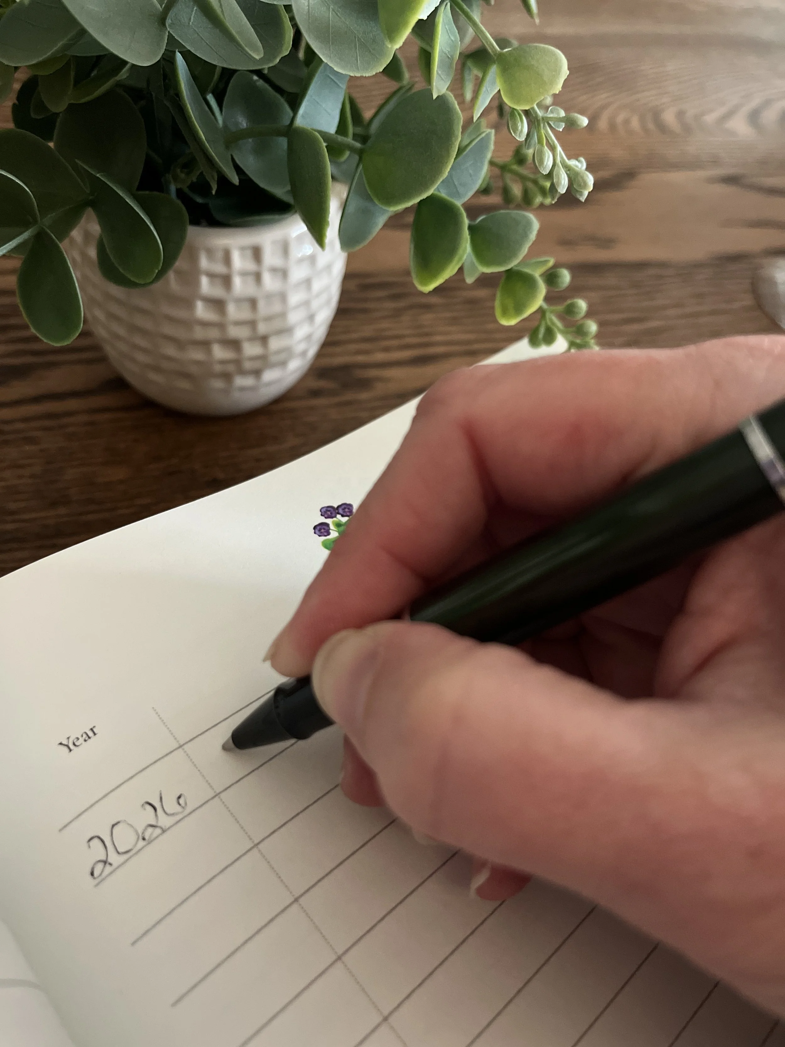 A person's hand holding a black pen writing '2026' on a page within the Keeping Book with a floral peaking out at the top of the page. In the background, there's a potted plant with green leaves and small buds on a wooden surface.