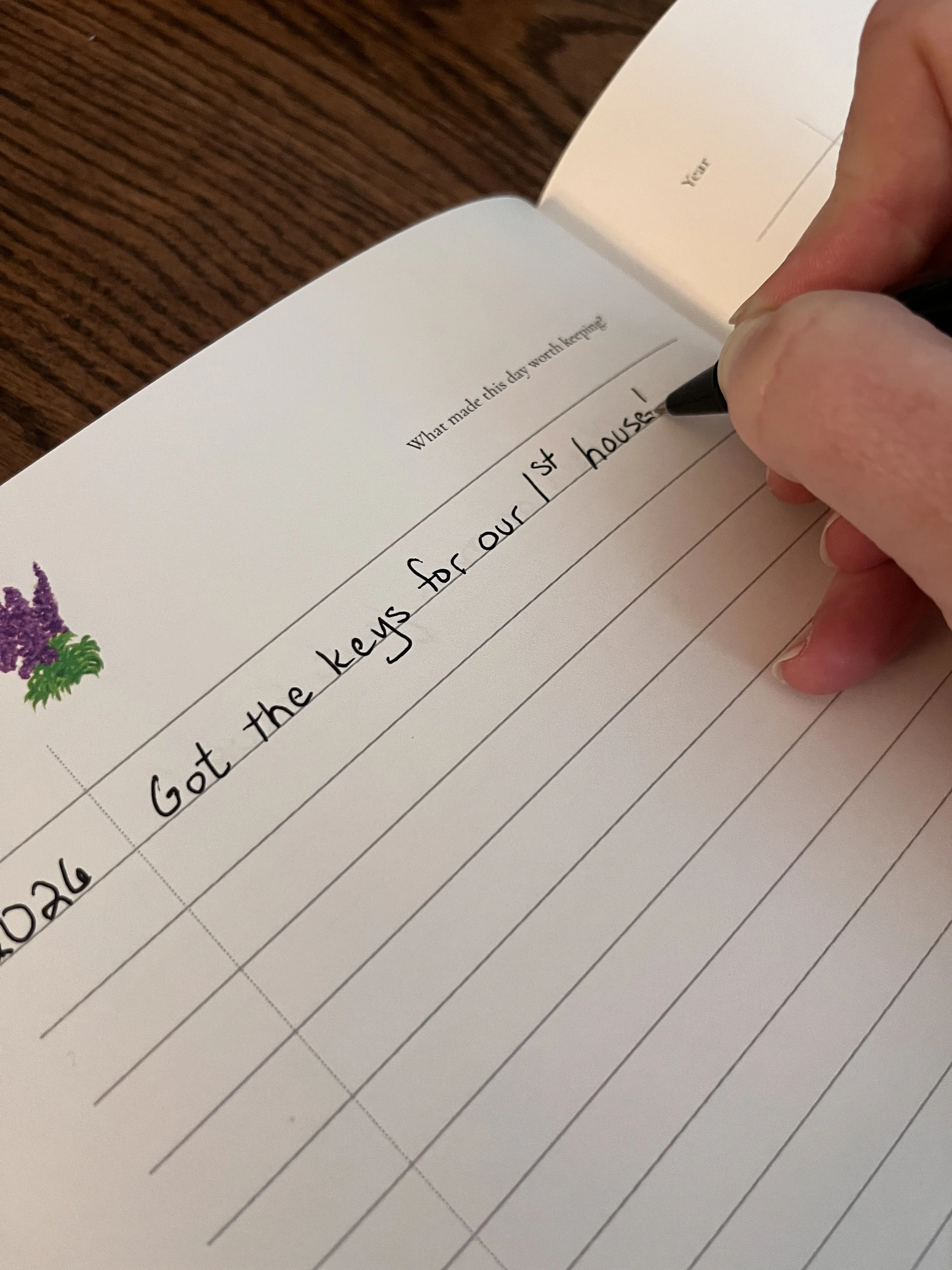 A person writing in a notebook with a black pen. The page contains a prompt 'What made this day worth keeping?' and the person has written '2026 - Got the keys for our 1st house!'.