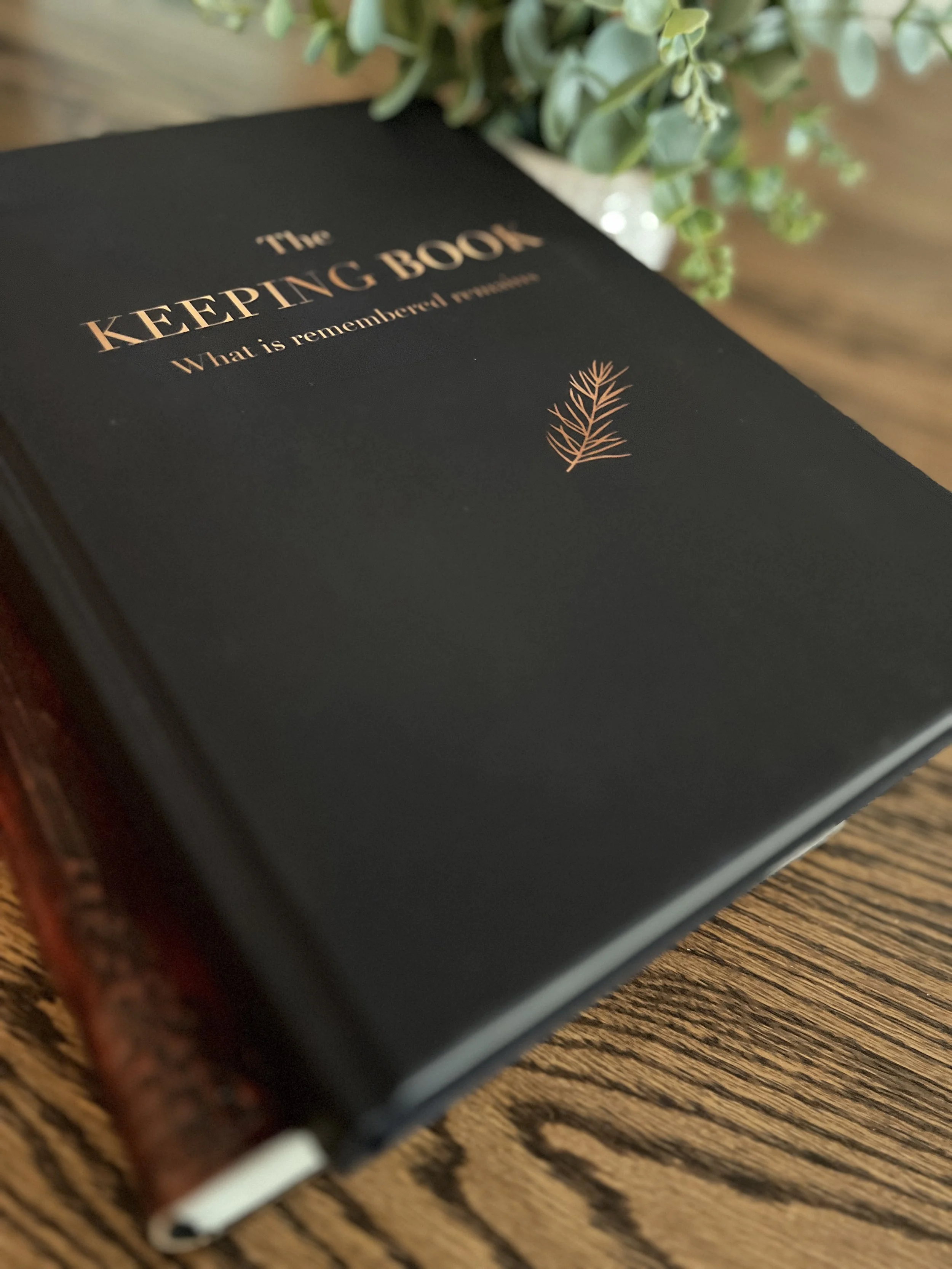 Close-up of a black book titled 'The Keeping Book' with a gold foil illustration of a sprig of rosemary. The book is on a wooden table next to a green eucalyptus plant in a white pot.