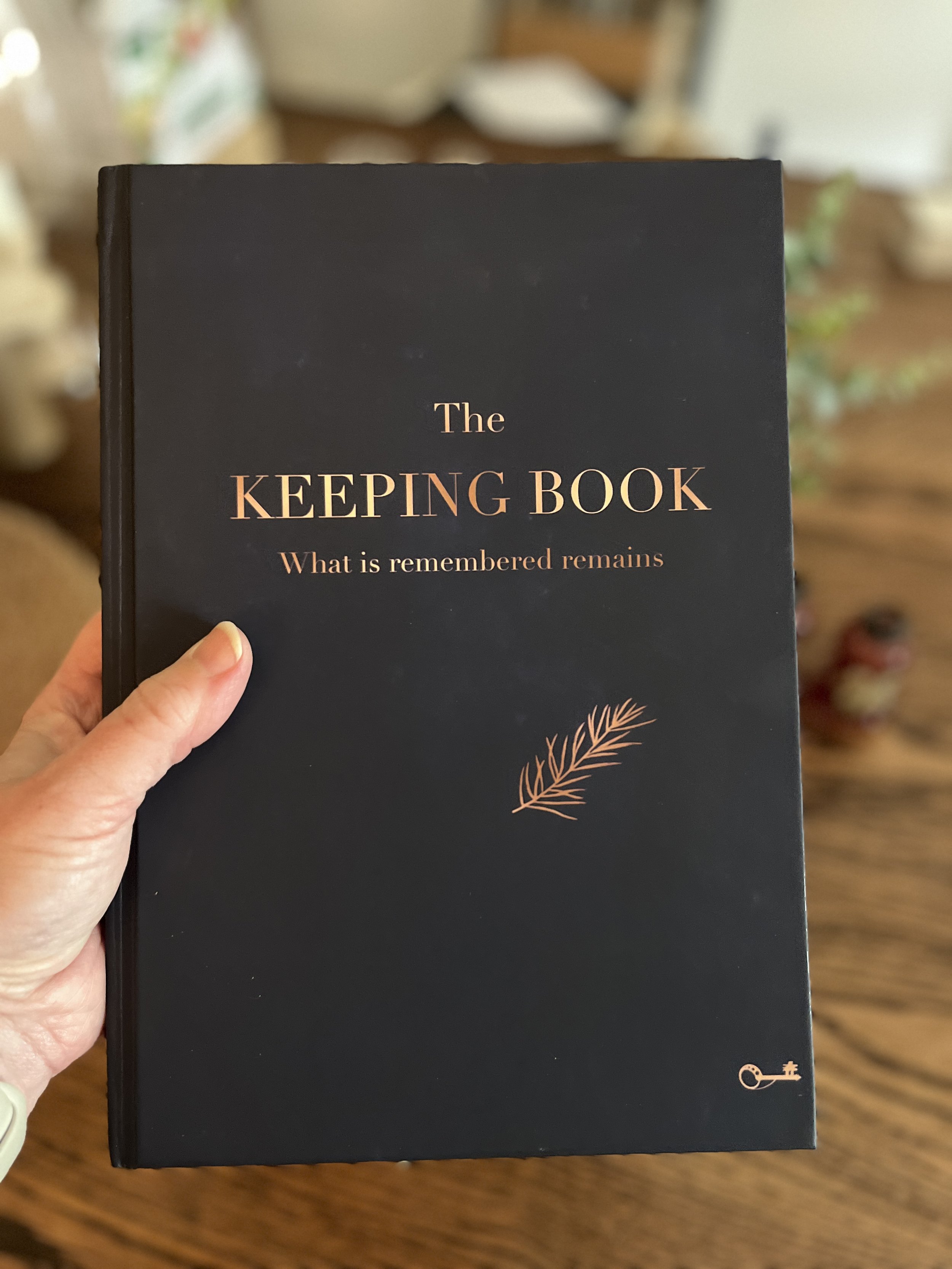A person holding a black book titled 'The Keeping Book' with the subtitle 'What is remembered remains' written in gold foil. There is a small decorative element of a sprig of rosemary and a key icon on the cover.