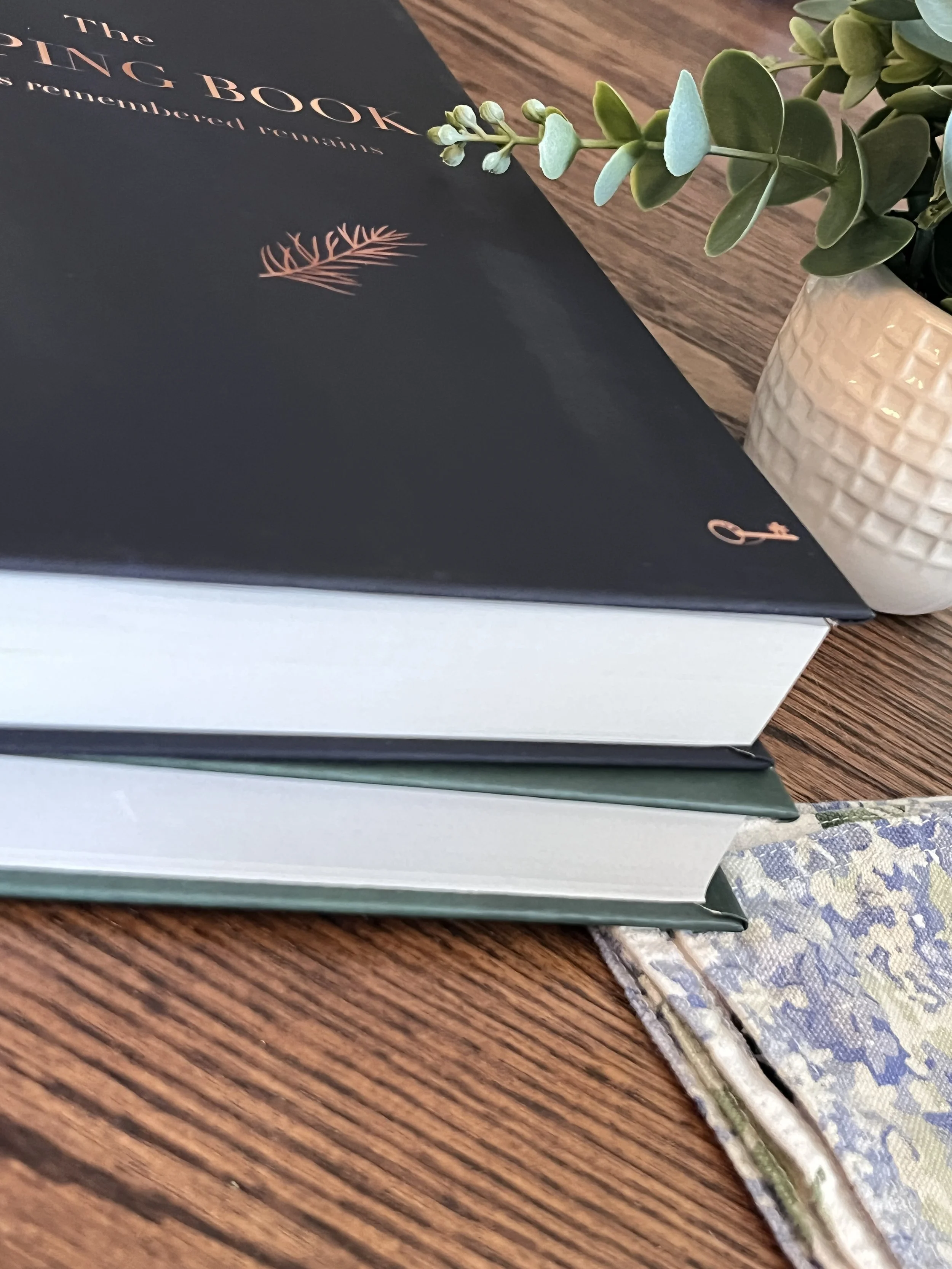 Navy and forest green Keeping Book memory journals stacked together beside eucalyptus leaves on a wooden table.