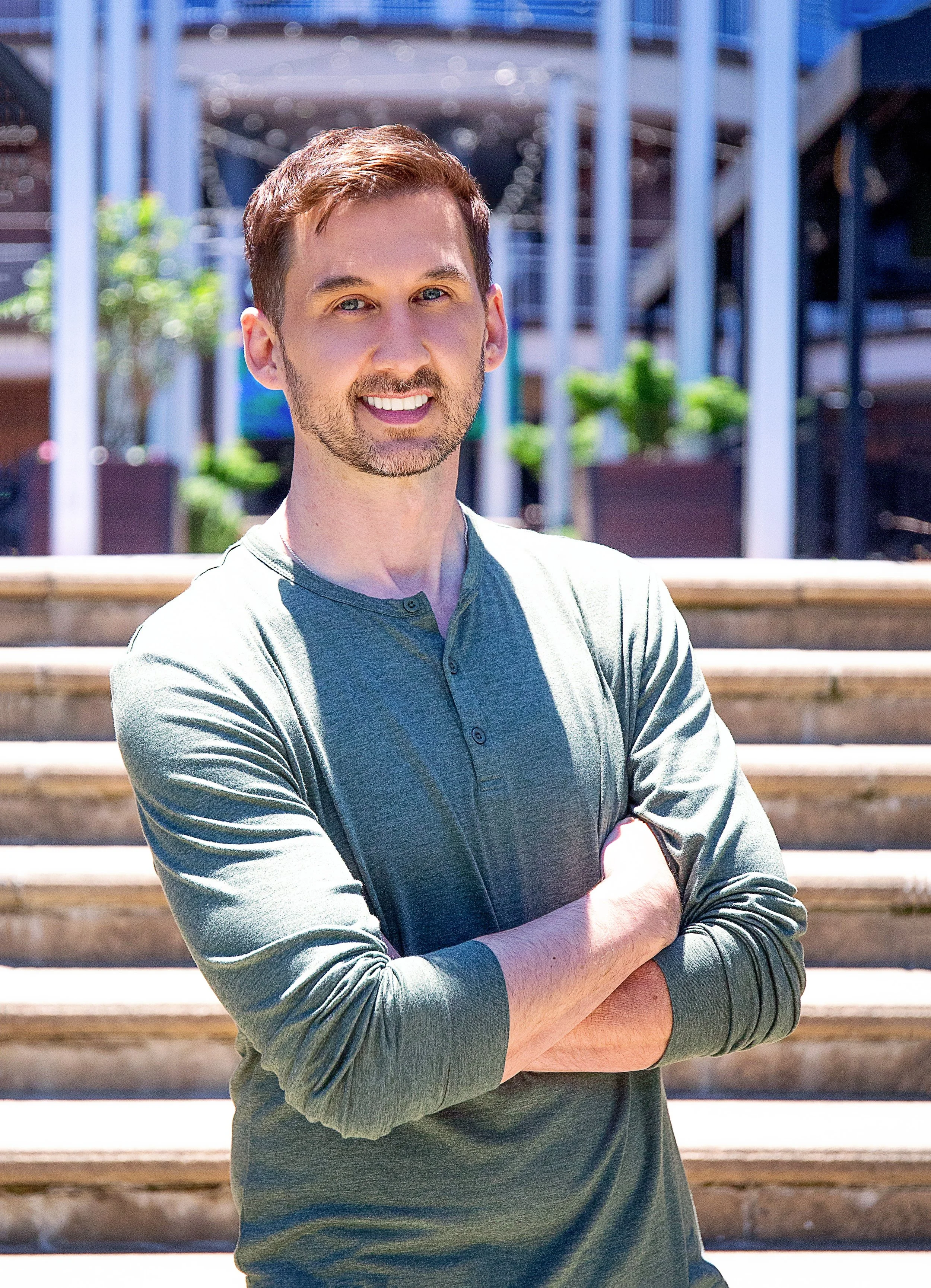 A young man with brown hair and a beard standing outdoors at the top of wooden stairs with arms crossed, smiling, wearing a long-sleeve gray shirt.